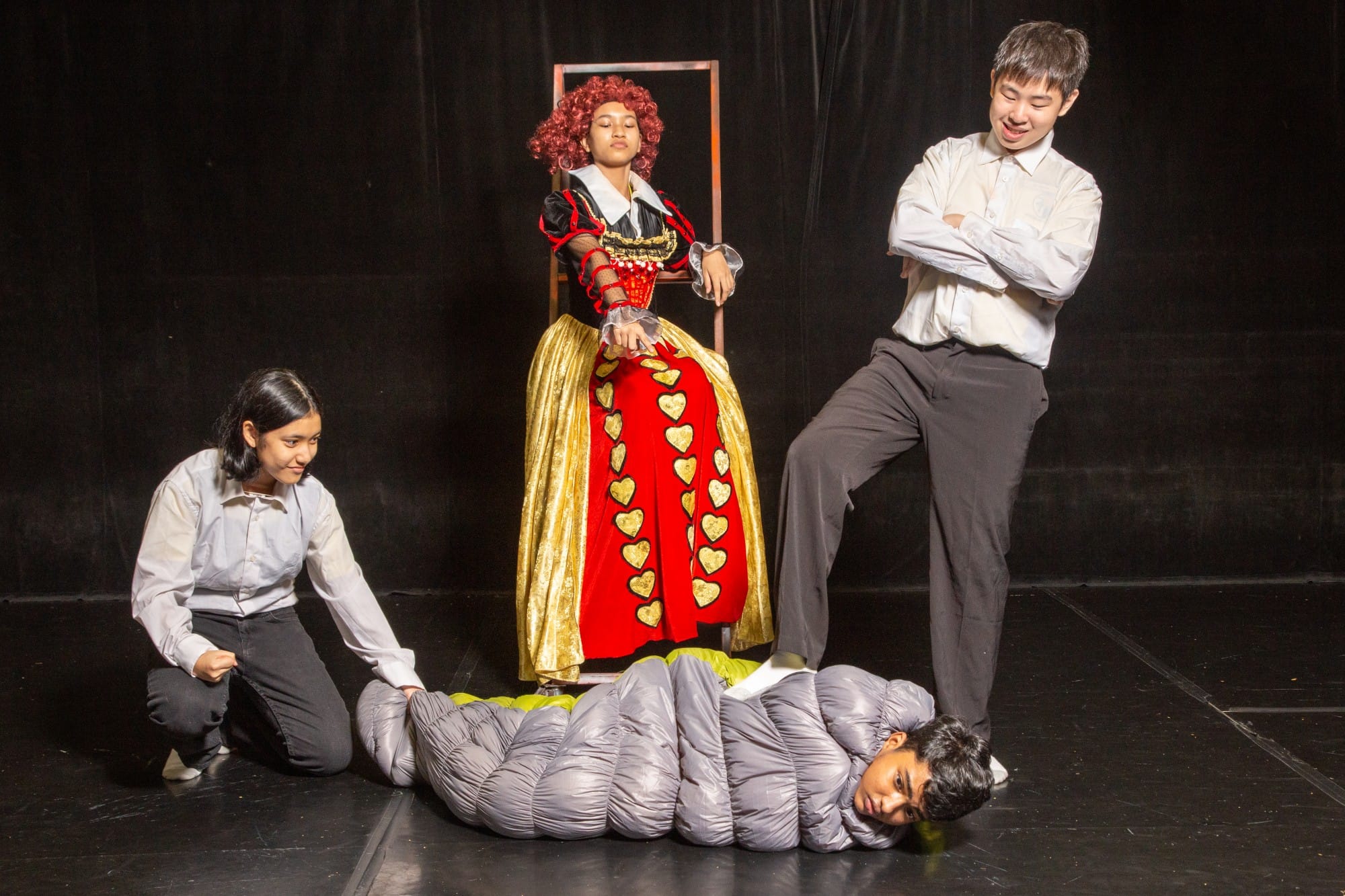 Four students perform a stage play; one wears a Queen of Hearts costume, one lies in a caterpillar suit on the floor.