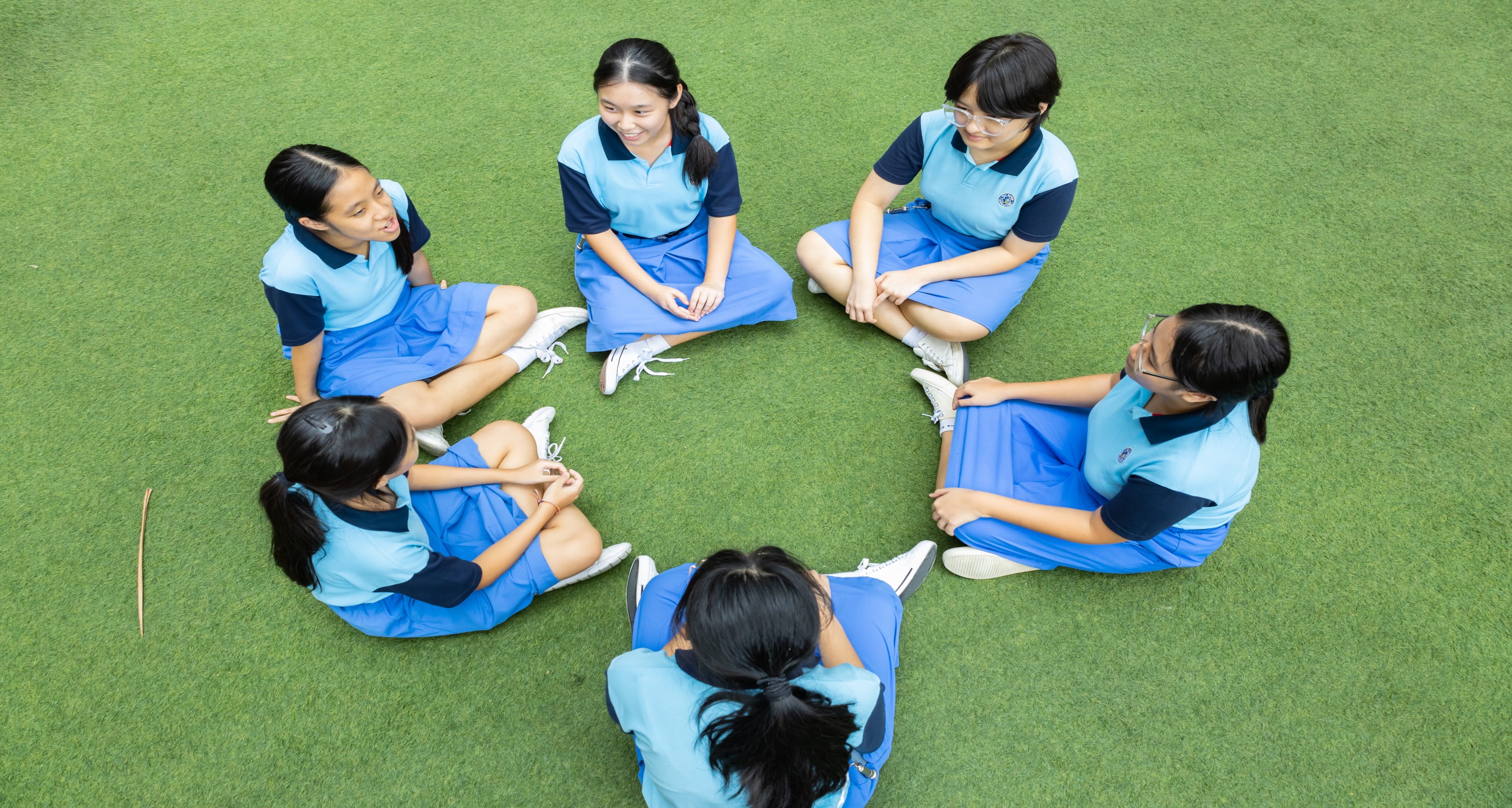 Six girls in blue school uniforms sit in a circle on green grass, talking and smiling.