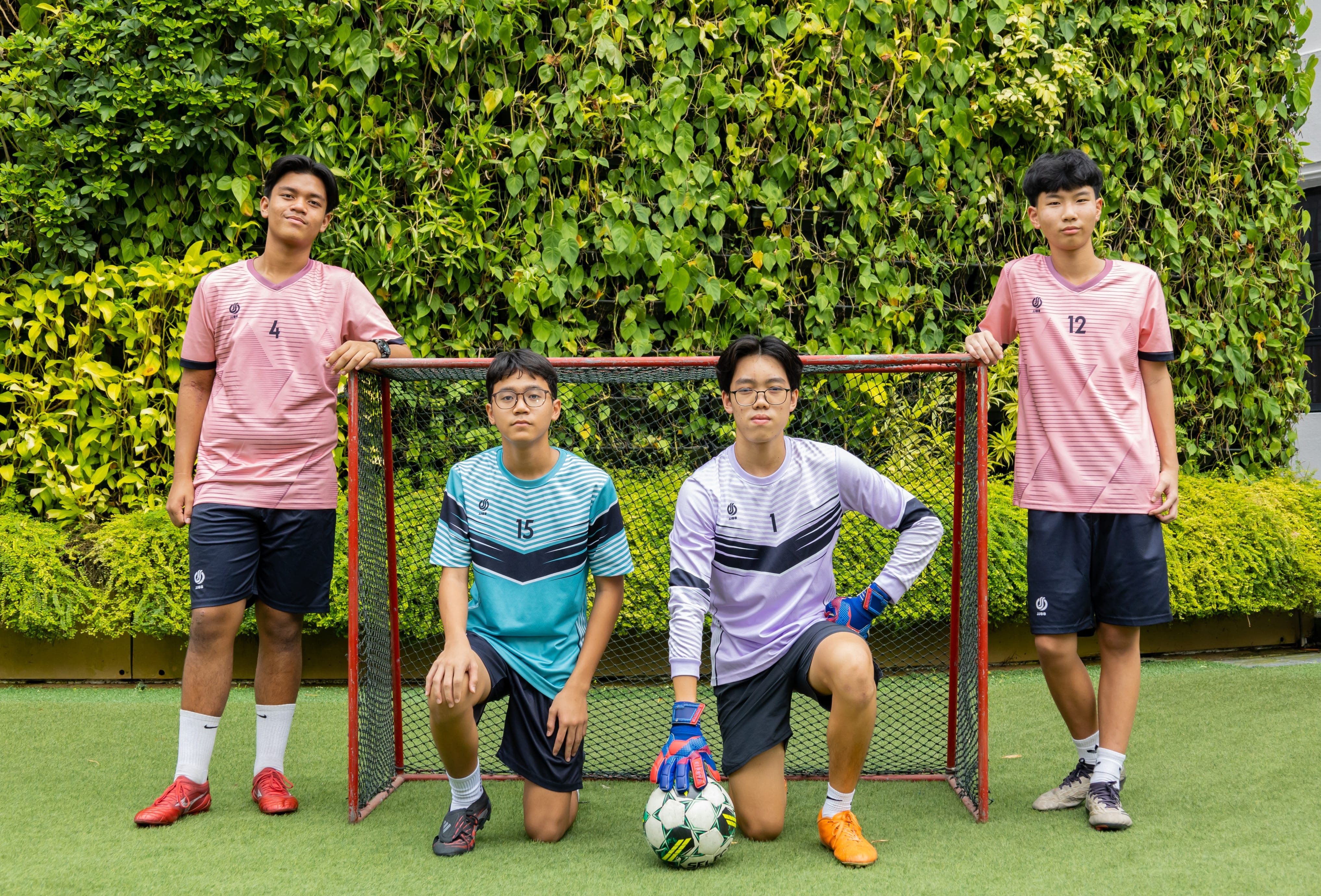 Four male students in football attire posing in front of a small goal on an artificial turf field, with a green hedge in the background.