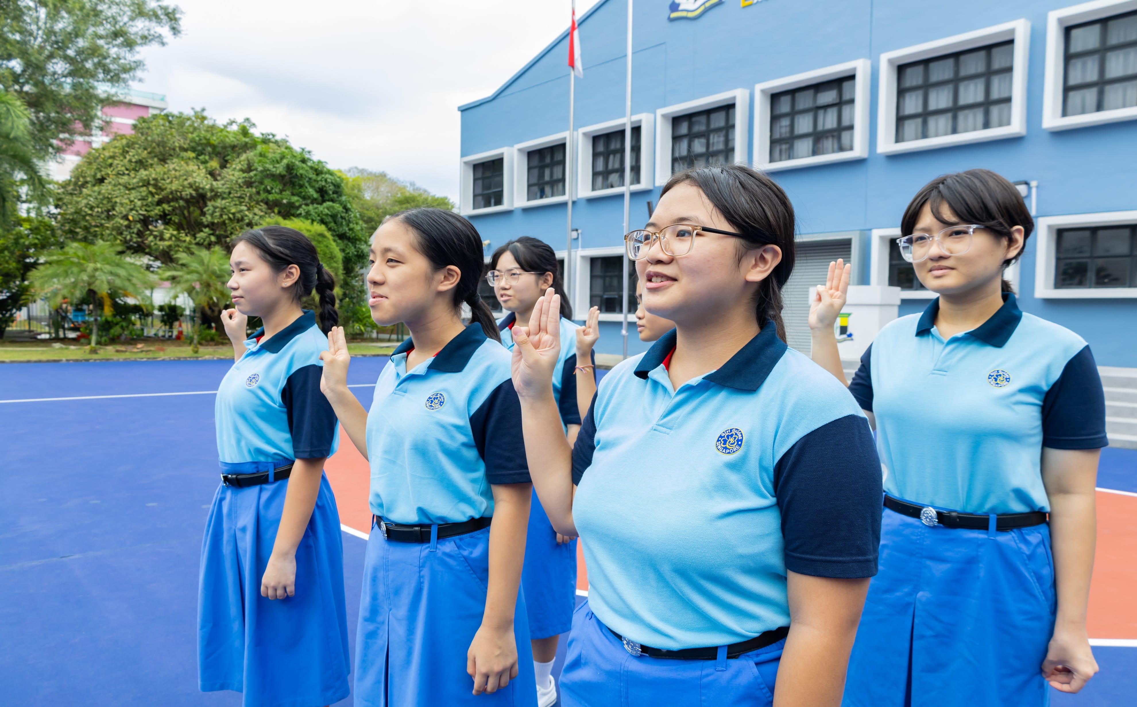 Five female students in blue school uniforms raise their right hands in a pledge on an outdoor court.
