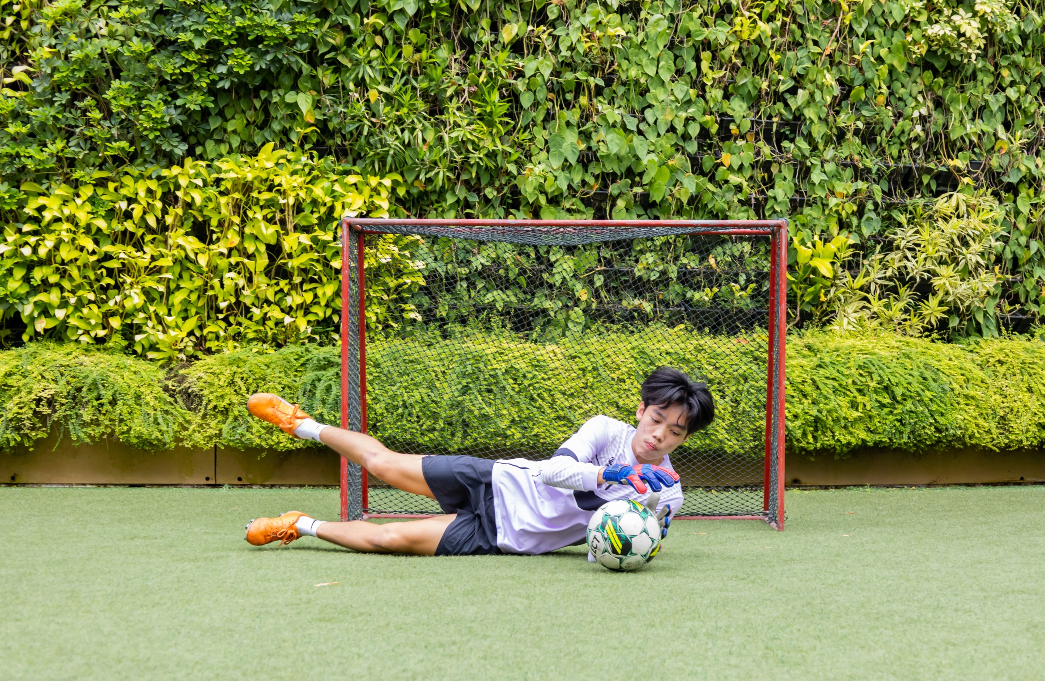 A young goalkeeper dives to save a soccer ball in front of a small red goal on green turf.