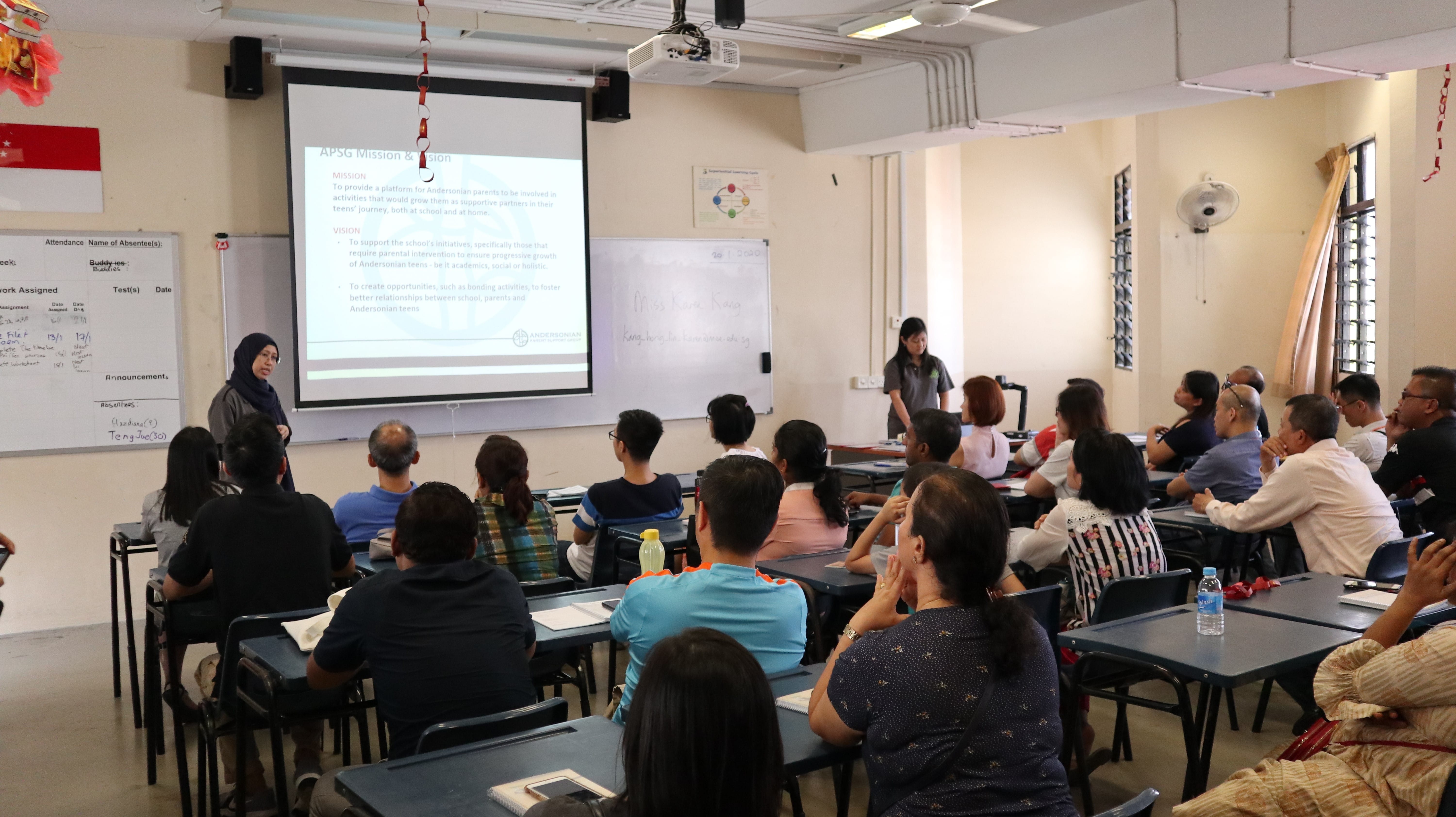 A presenter stands beside a projected slide titled APSG Mission and Vision while adults sit at desks in a classroom.