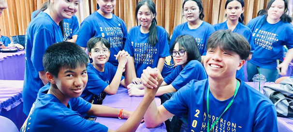 Two boys arm wrestling at a table while a group of smiling teens in matching blue shirts watch.