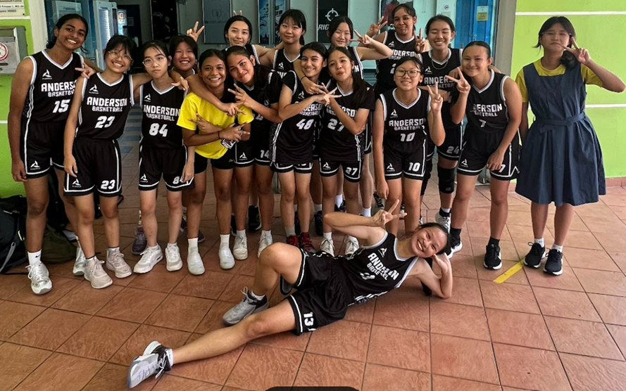 A girls basketball team in black Anderson uniforms poses together, flashing peace signs, with one player lying on the floor.