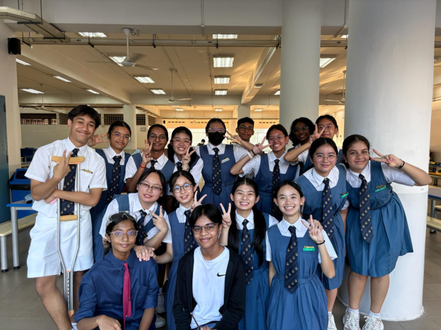 Group of school students in blue and white uniforms smiling and posing with peace signs indoors.