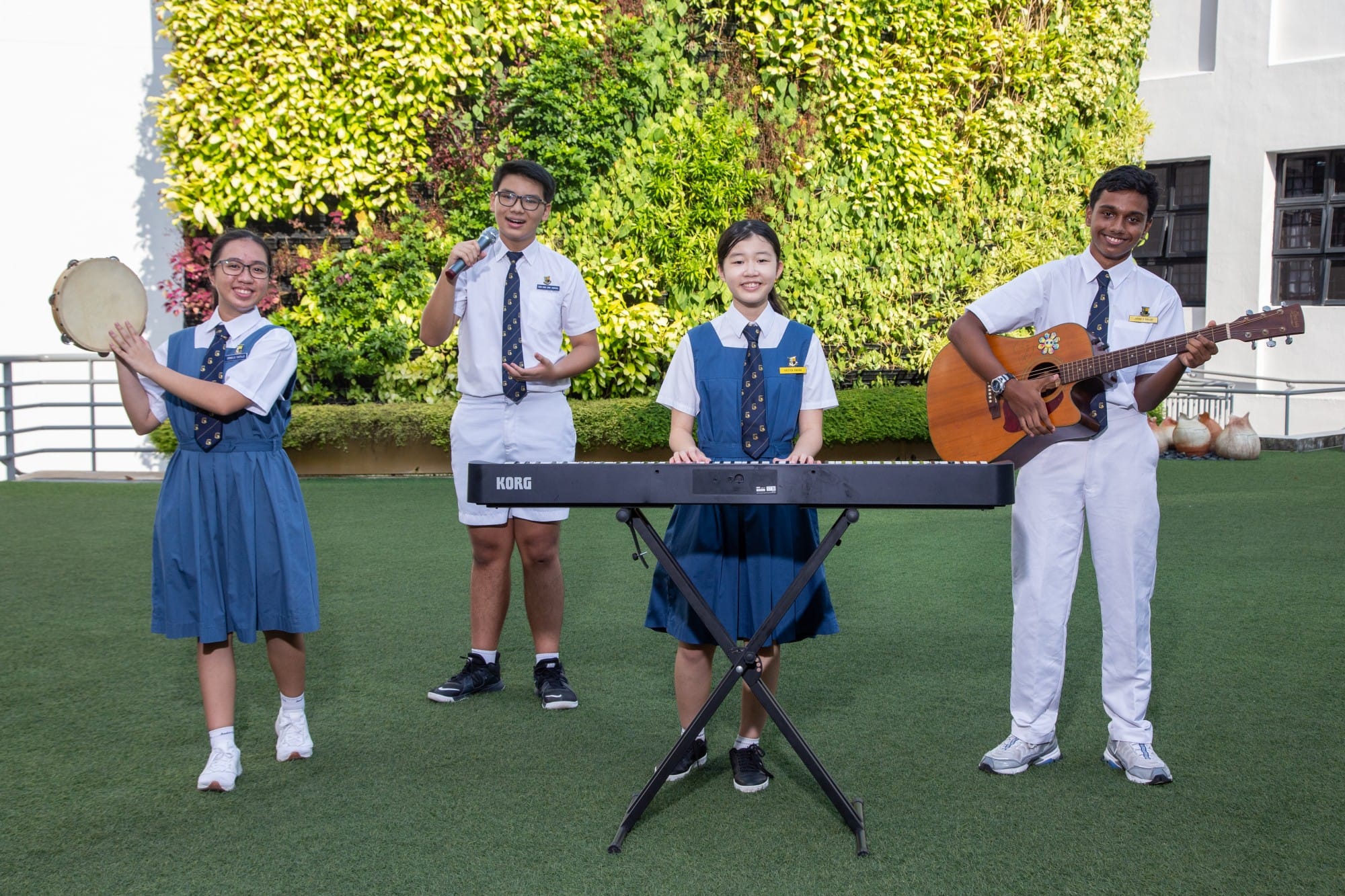 Four uniformed students perform outdoors with tambourine, microphone, keyboard, and guitar in front of a green plant wall.