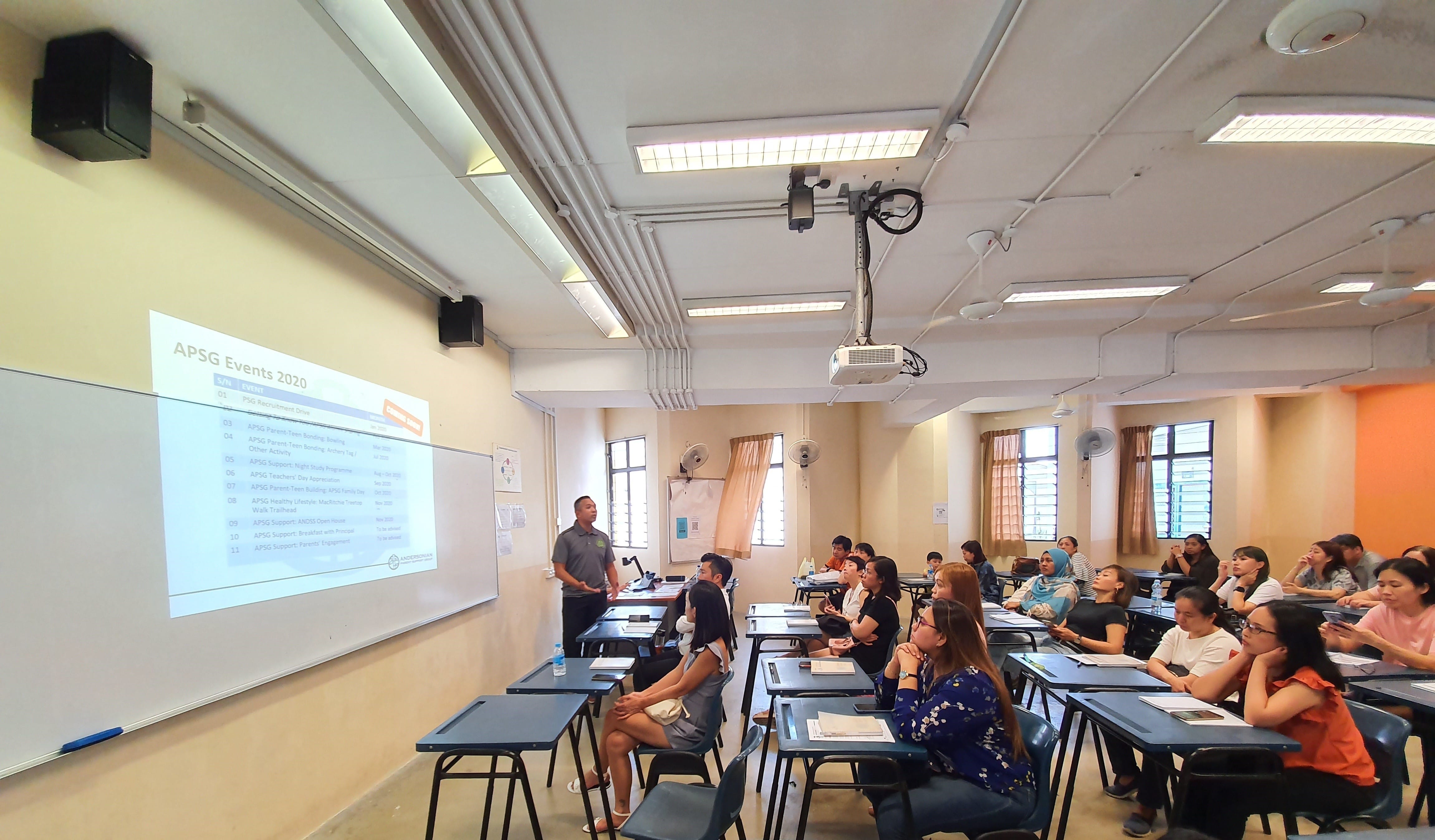 A man presents an APSG Events 2020 slideshow to a room full of seated attendees in a classroom.