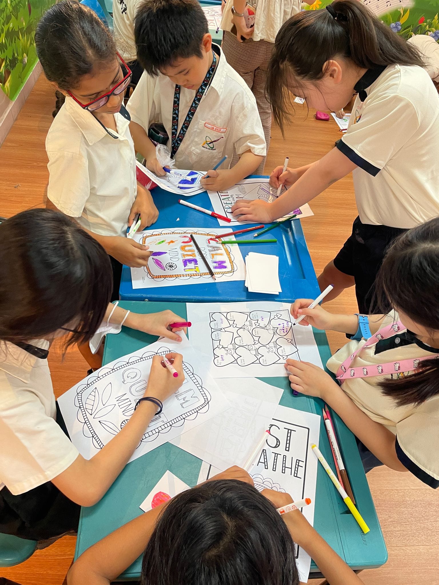 Five children in school uniforms sit around a blue table, coloring worksheets with markers and pencils.