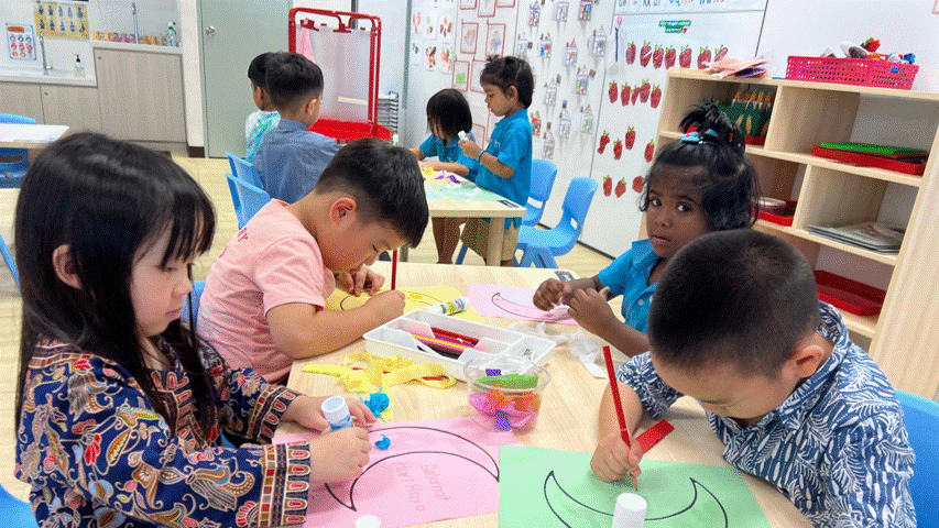 Several young children sit at tables doing arts and crafts activities in a colorful classroom.
