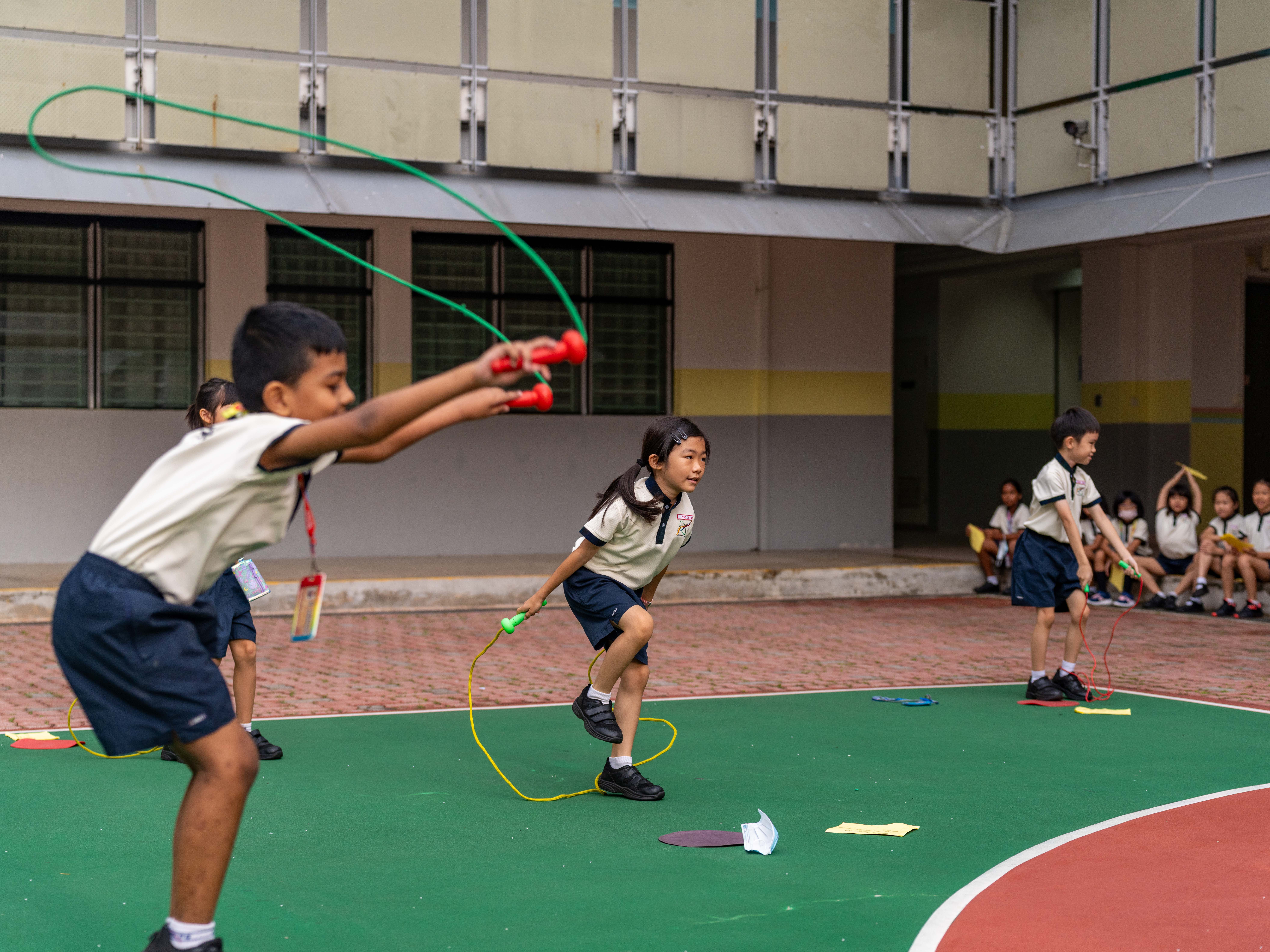 Schoolchildren skipping rope on a playground, with a boy swinging a green rope in the foreground.