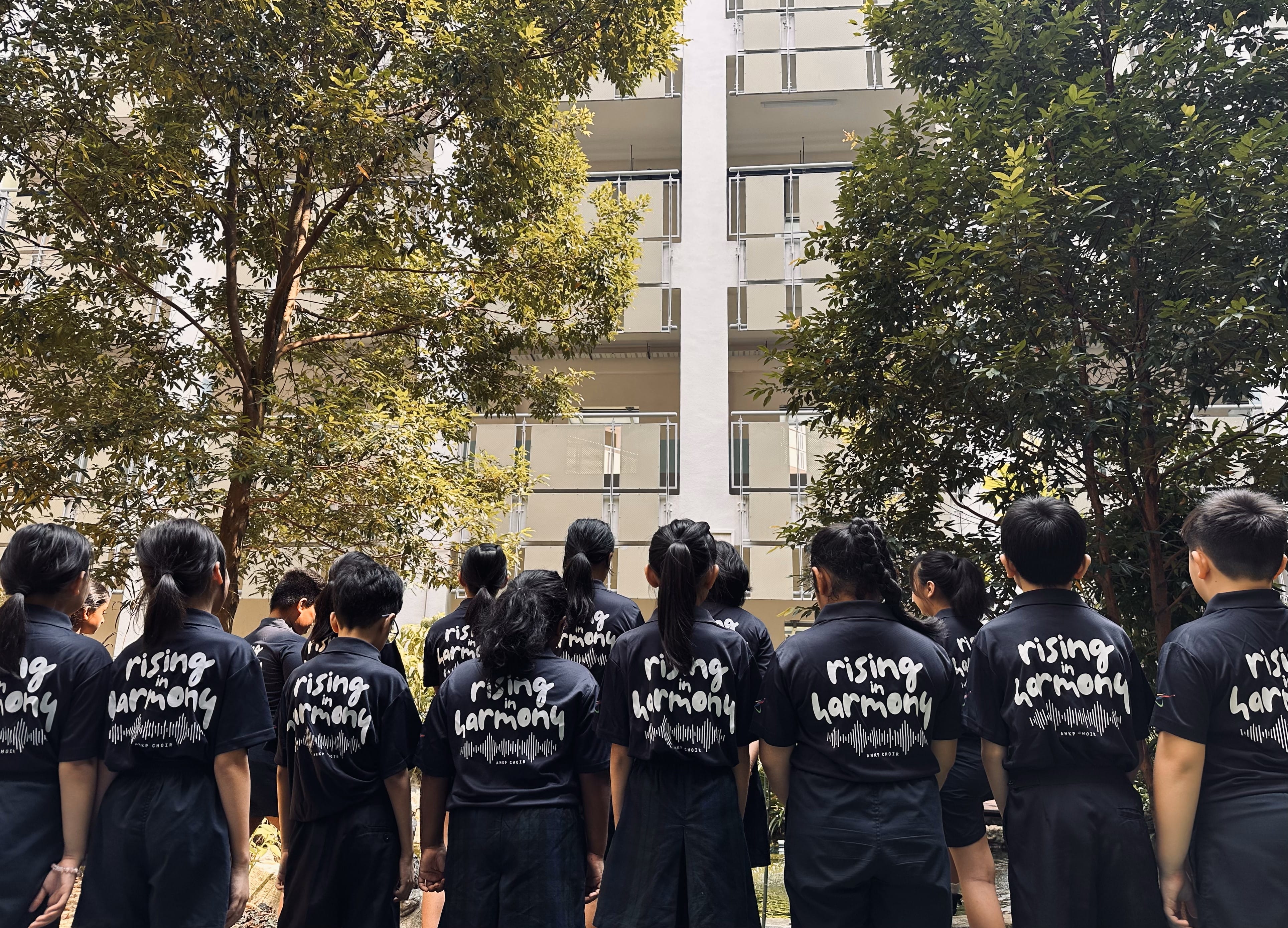 Group of young people in black shirts reading "rising in harmony" stand facing a multi-story building outdoors.