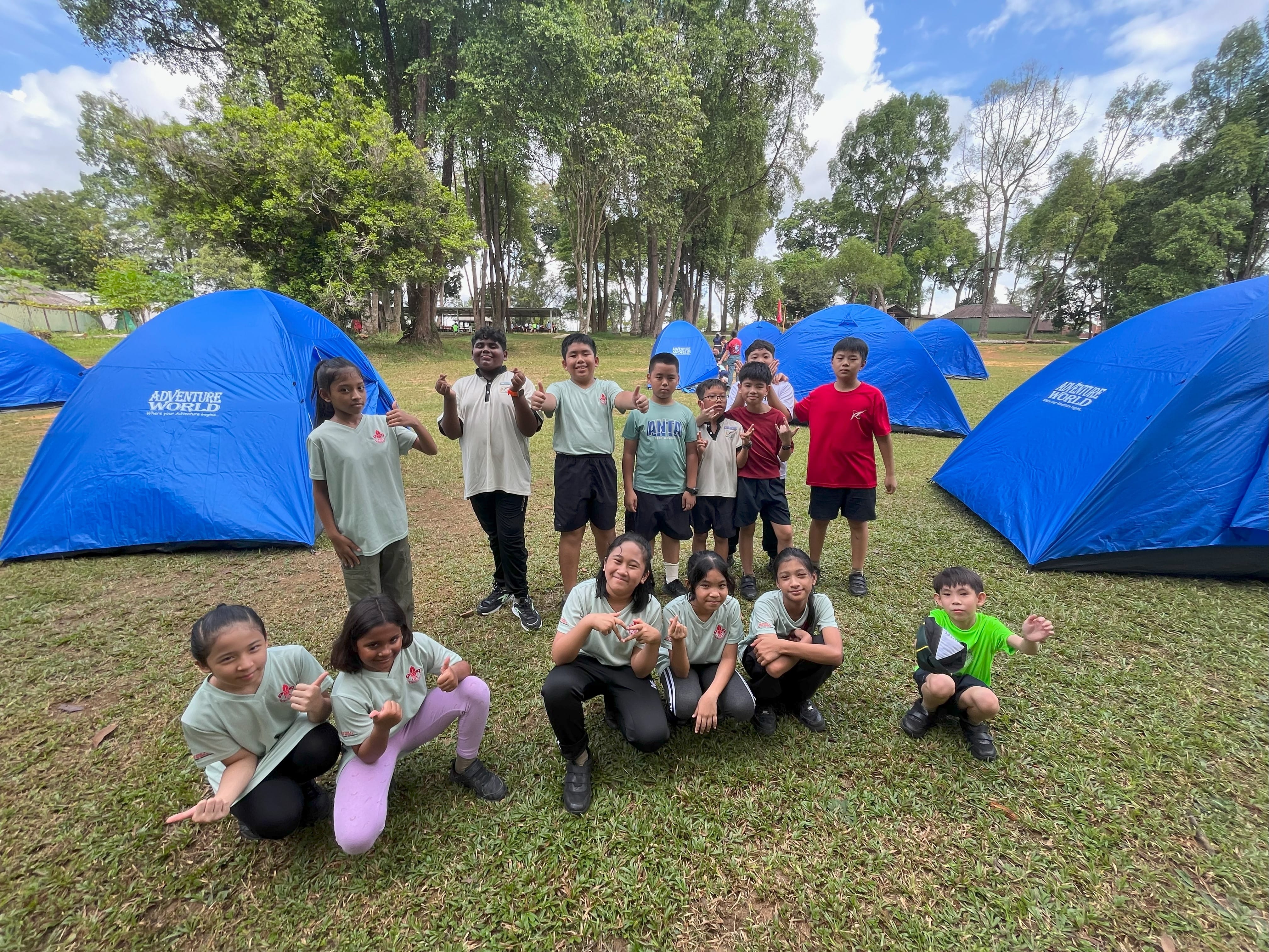 Group of kids posing on grass at a campsite with several bright blue dome tents and tall trees behind.