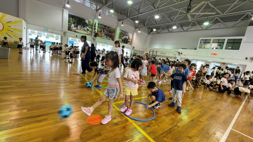 Children play with hoops and soccer balls on a wooden gymnasium floor during a school physical education session.