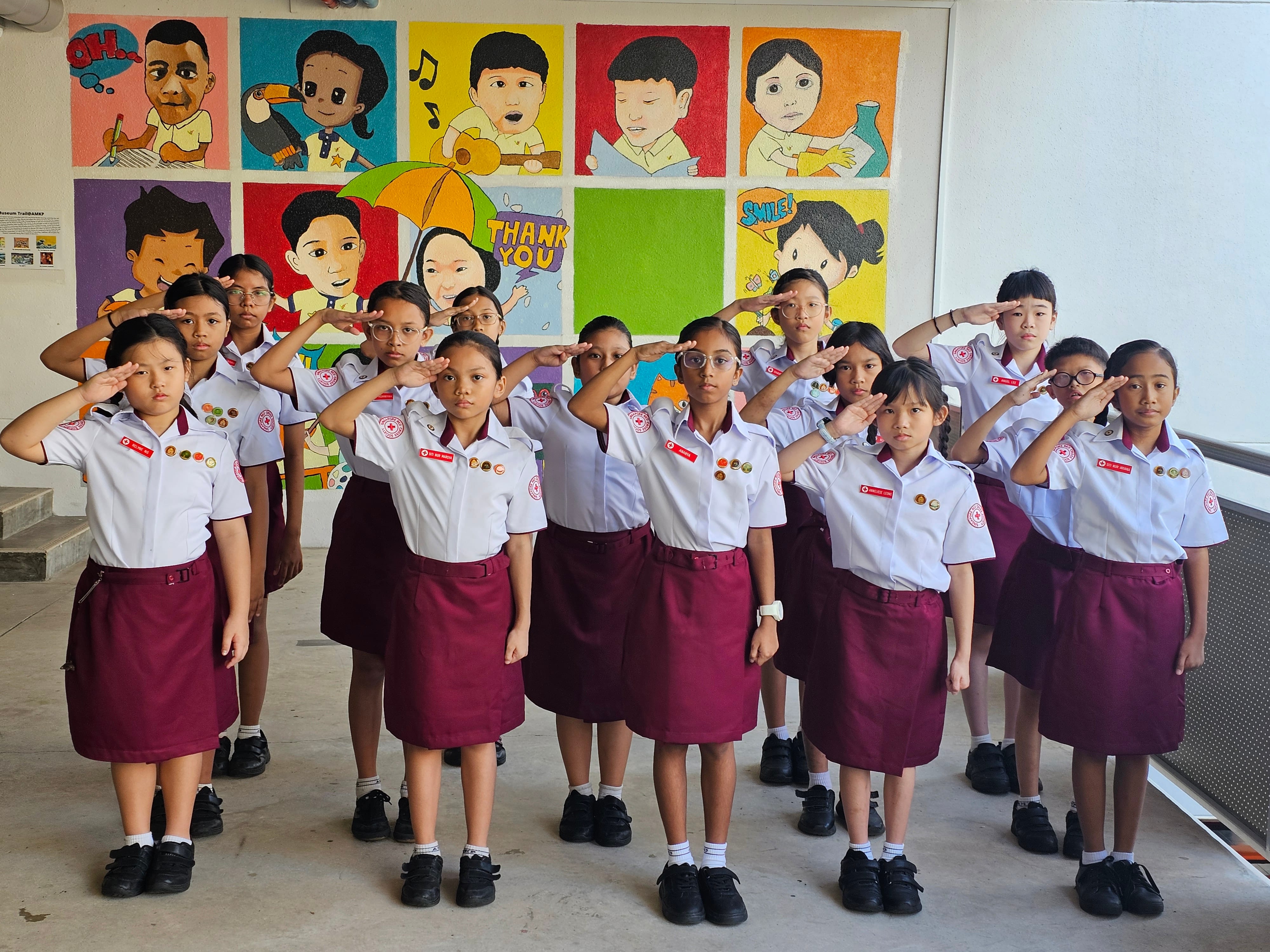 A group of schoolgirls in white shirts and maroon skirts saluting in front of a colorful cartoon mural.