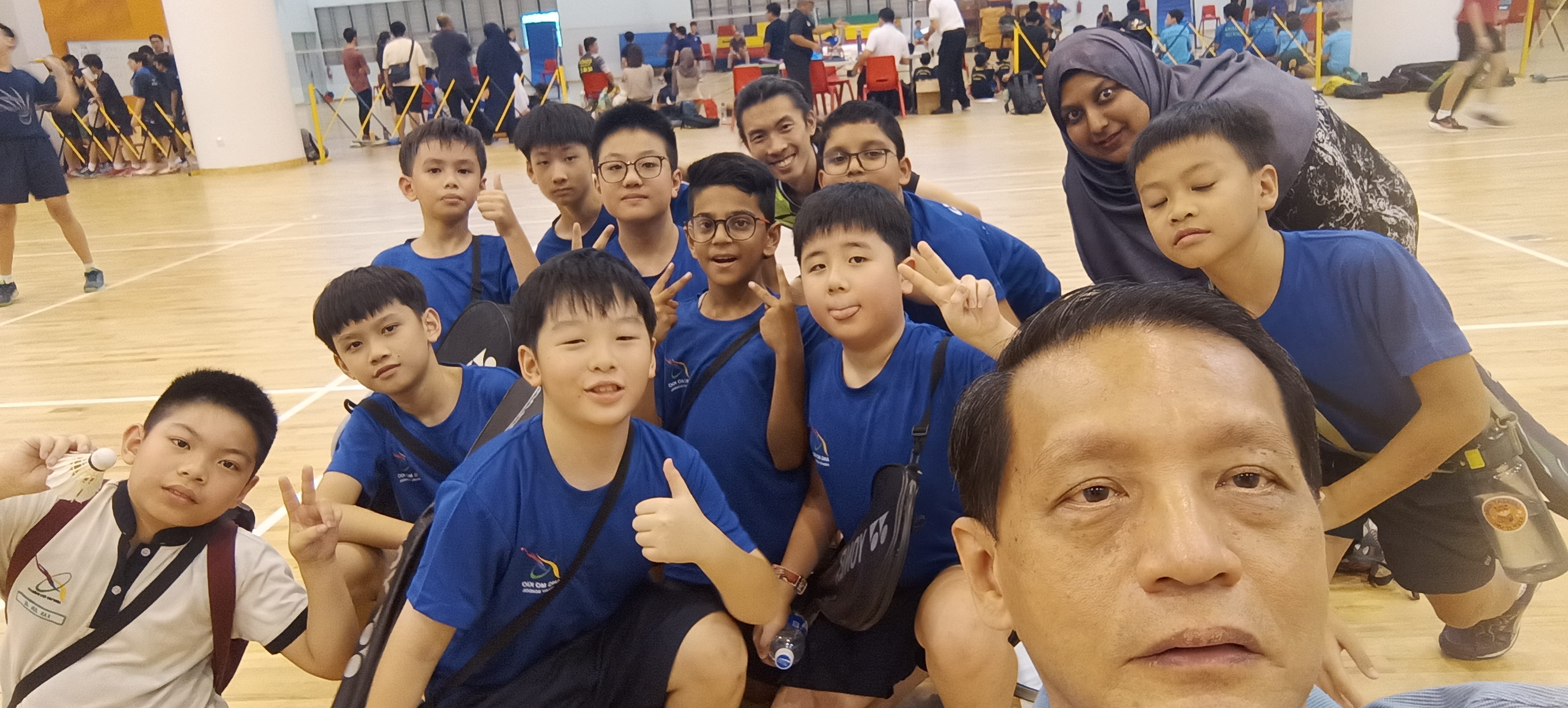 A group of children in blue shirts pose for a selfie with two adults in an indoor badminton sports hall.