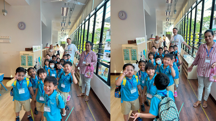 A group of young children in blue uniforms pose with adults in a bright indoor corridor with large windows.