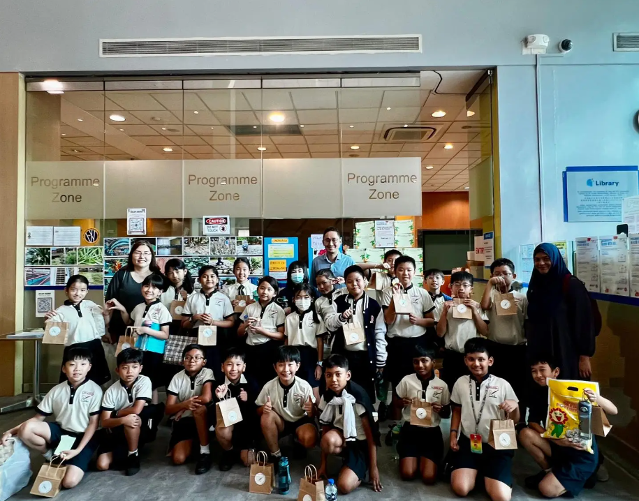 A large group of school children and teachers pose together in front of glass doors labeled Programme Zone, holding small paper bags.