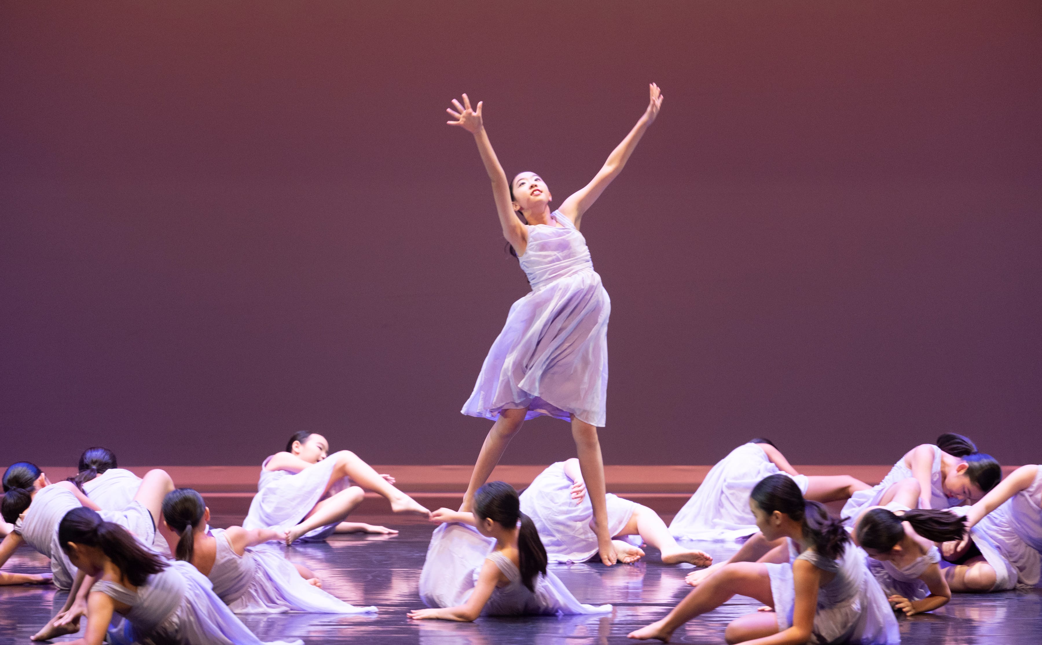 Ballet dancer in pale dress stands center with arms raised as others in similar dresses lie on stage floor.