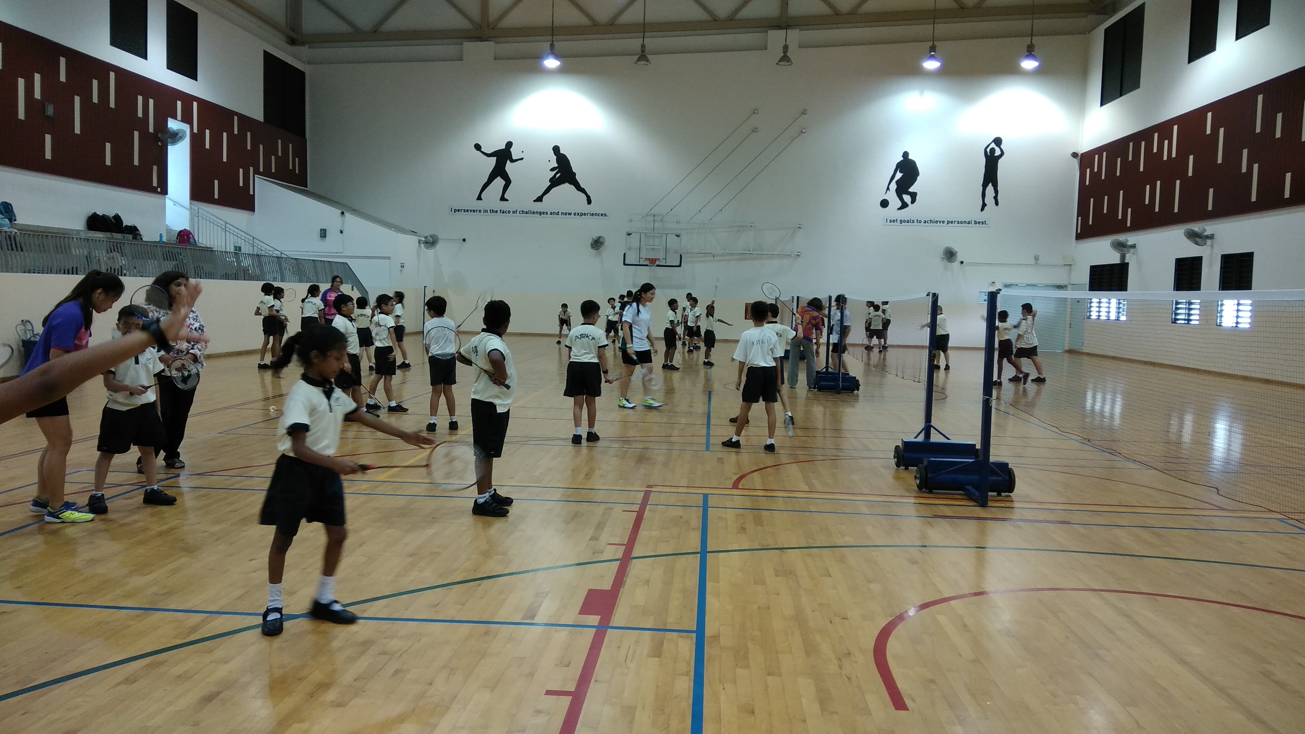 Children in school uniforms practice badminton with rackets in an indoor gymnasium with sport murals on the wall.