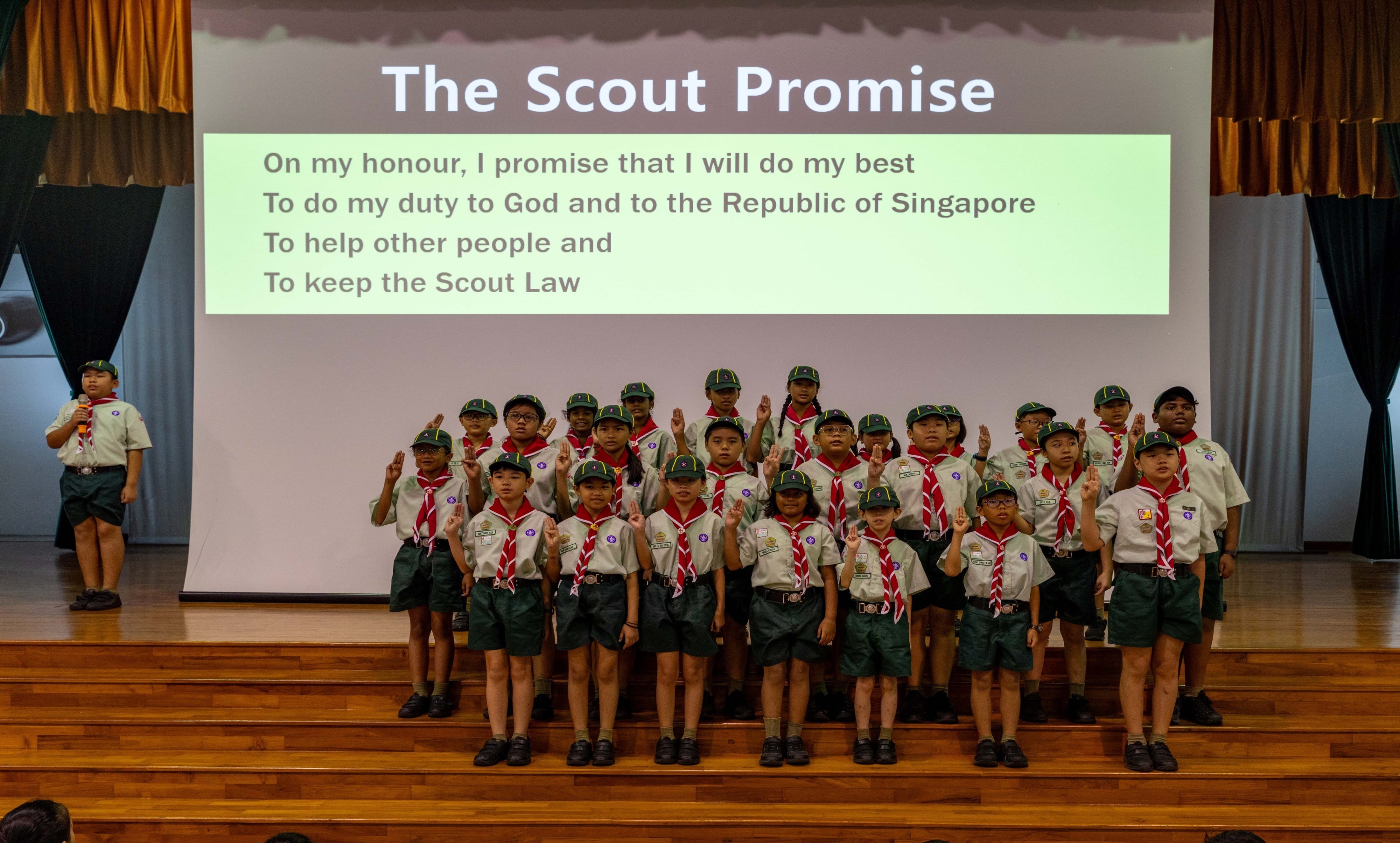 Children in Scout uniforms stand on a stage raising three fingers, reciting the Scout Promise shown on a screen behind them.