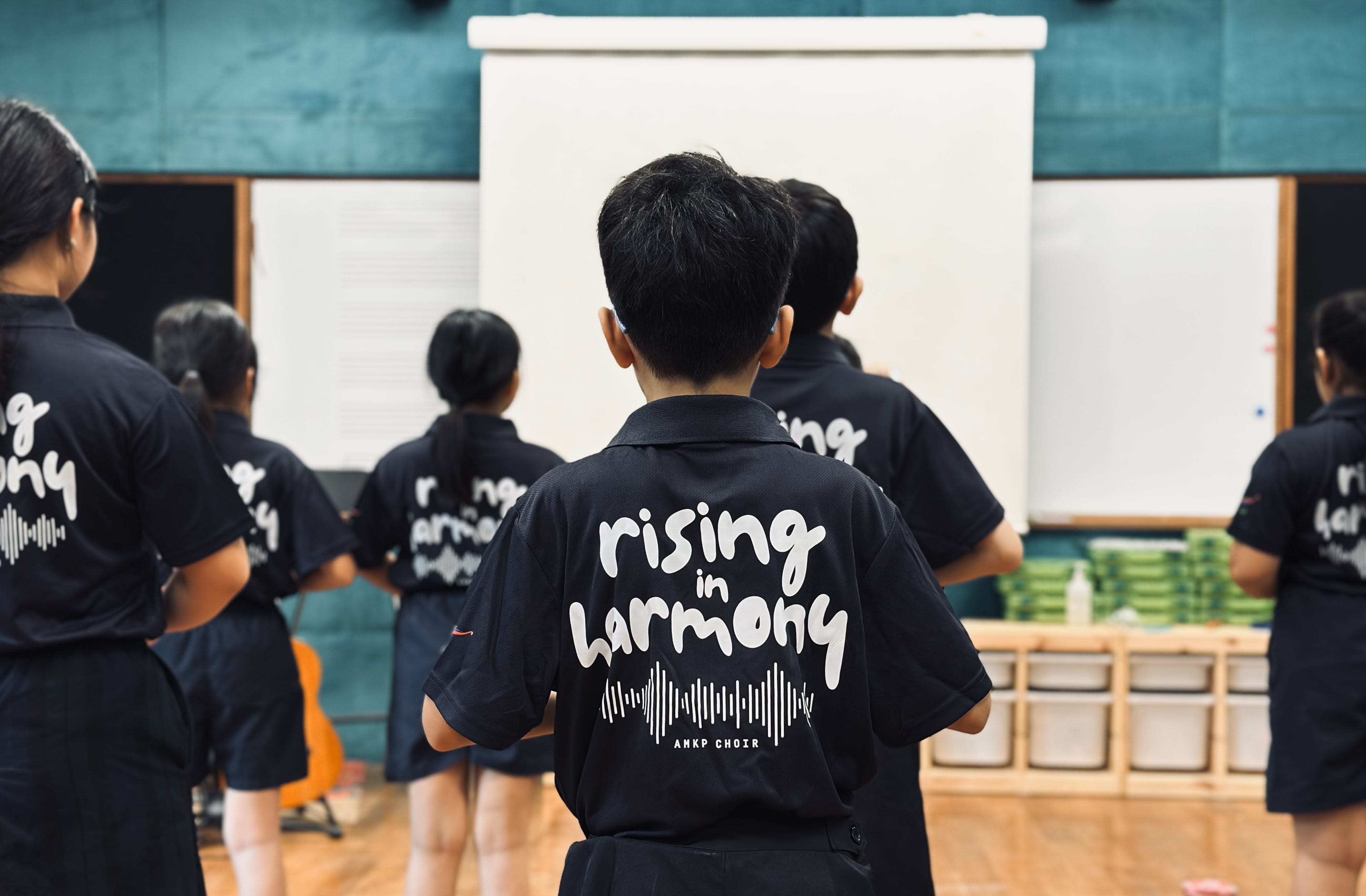 Children wearing black ANKP Choir shirts reading "rising in harmony" stand facing a whiteboard in a hall.