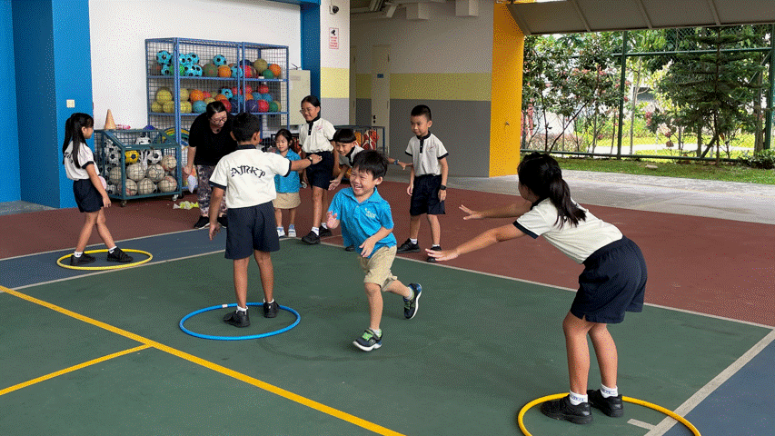 Children in school uniforms play an active game with hula hoops on a colorful outdoor court supervised by a teacher.