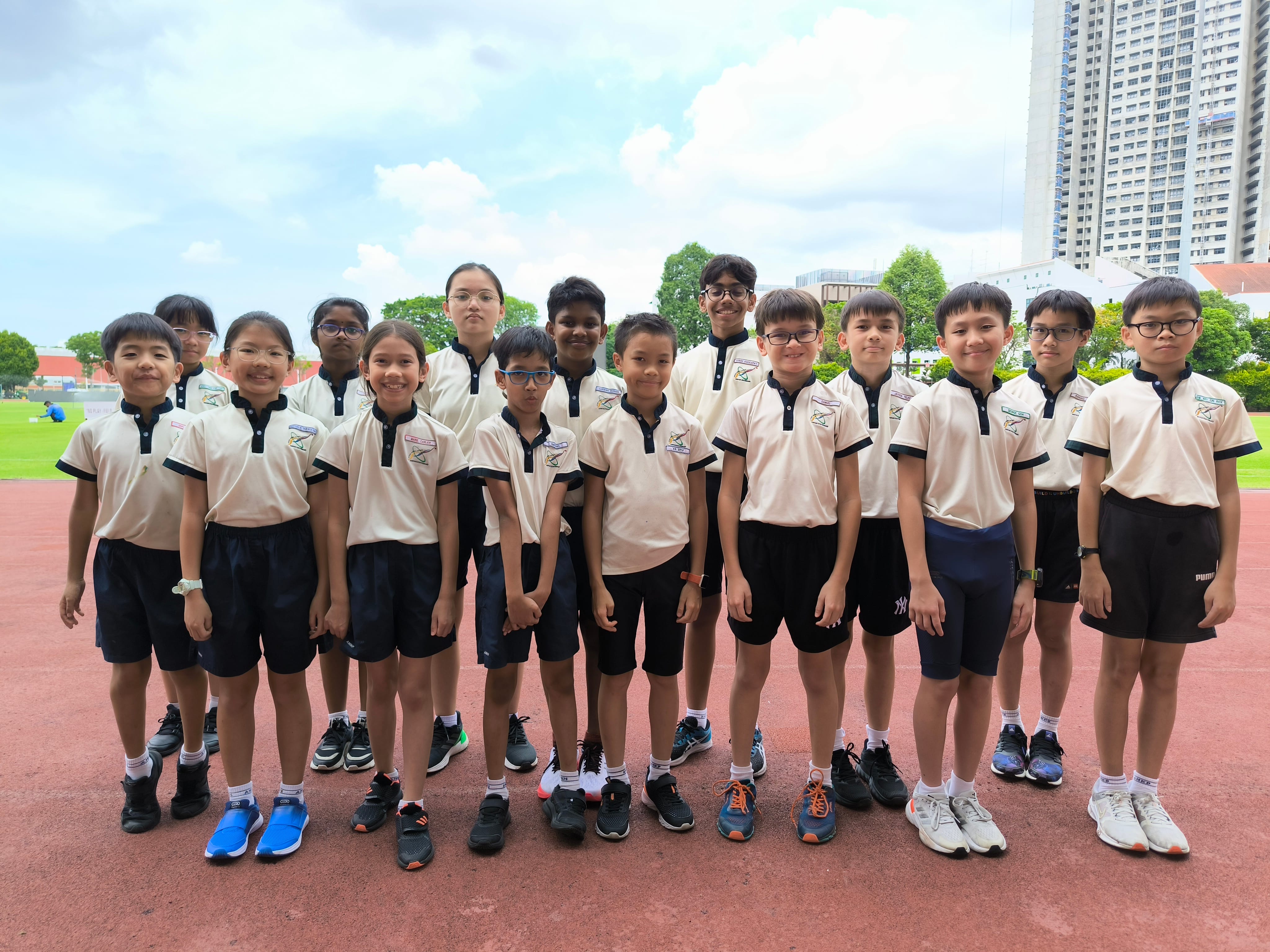A group of about 16 children in matching cream and black school uniforms posing on a red running track outdoors.