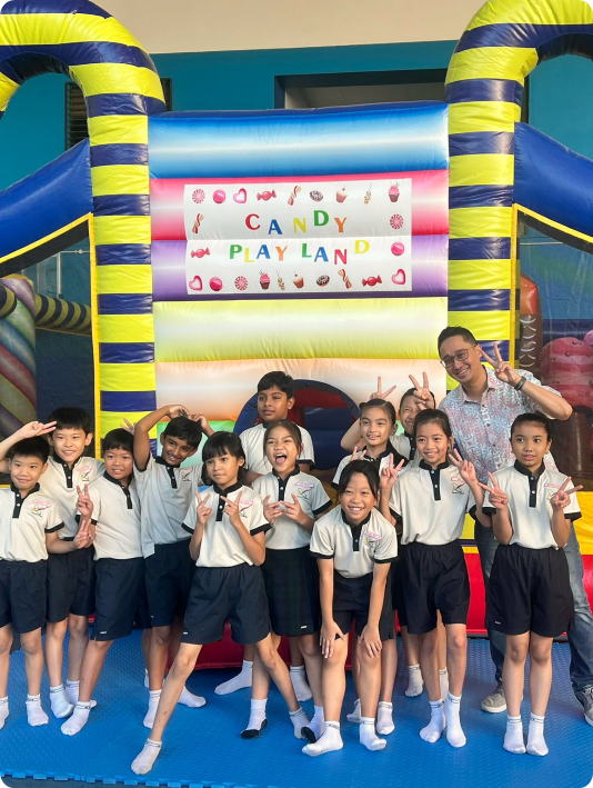 A group of school children and a teacher pose in front of a colorful inflatable bounce house labeled Candy Play Land.