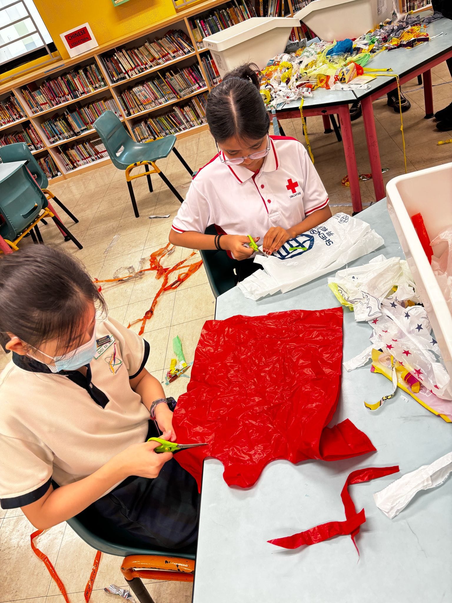 Two students in a school library cutting plastic bags with scissors at a table covered in colorful plastic pieces.