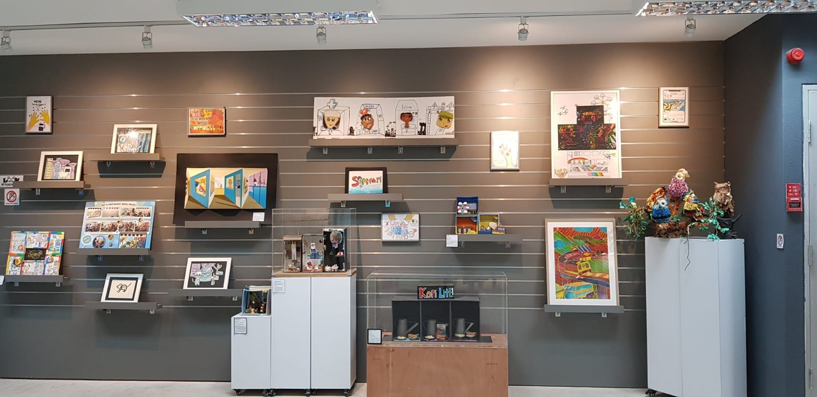 A gallery wall with multiple colorful artworks on shelves and display cases, lit by overhead track lighting.