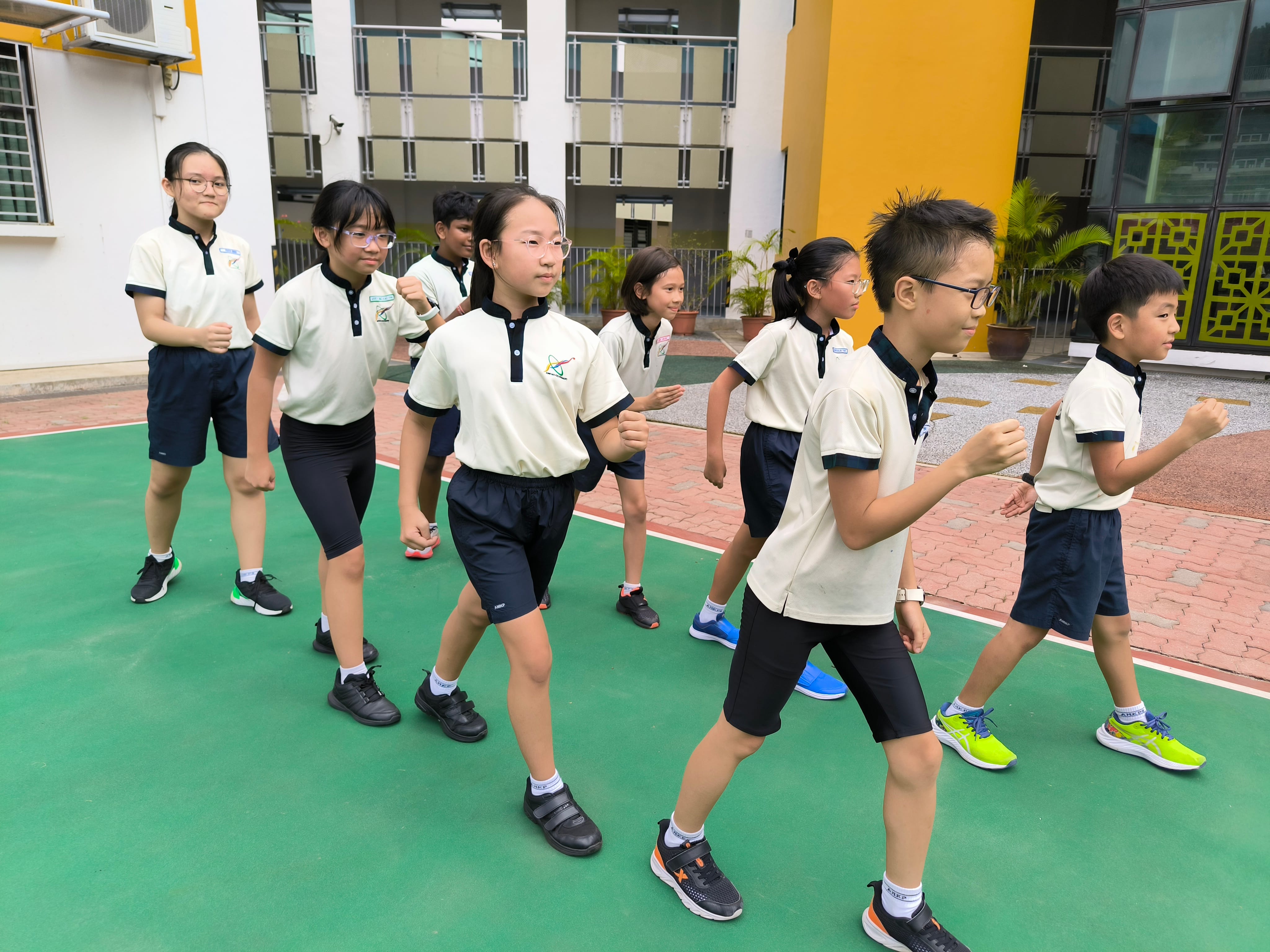 Eight primary school students in white and navy uniforms march in formation on a green outdoor court.