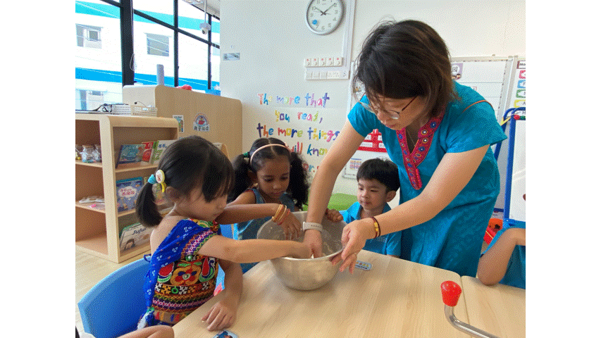 A teacher in teal helps three young children mix ingredients in a bowl at a classroom table.