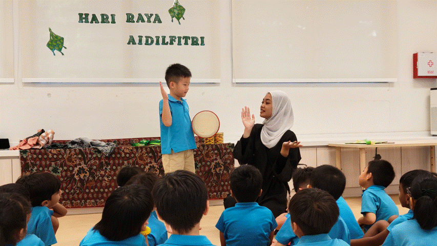 A woman in a hijab and a young boy stand before seated children under a Hari Raya Aidilfitri banner.