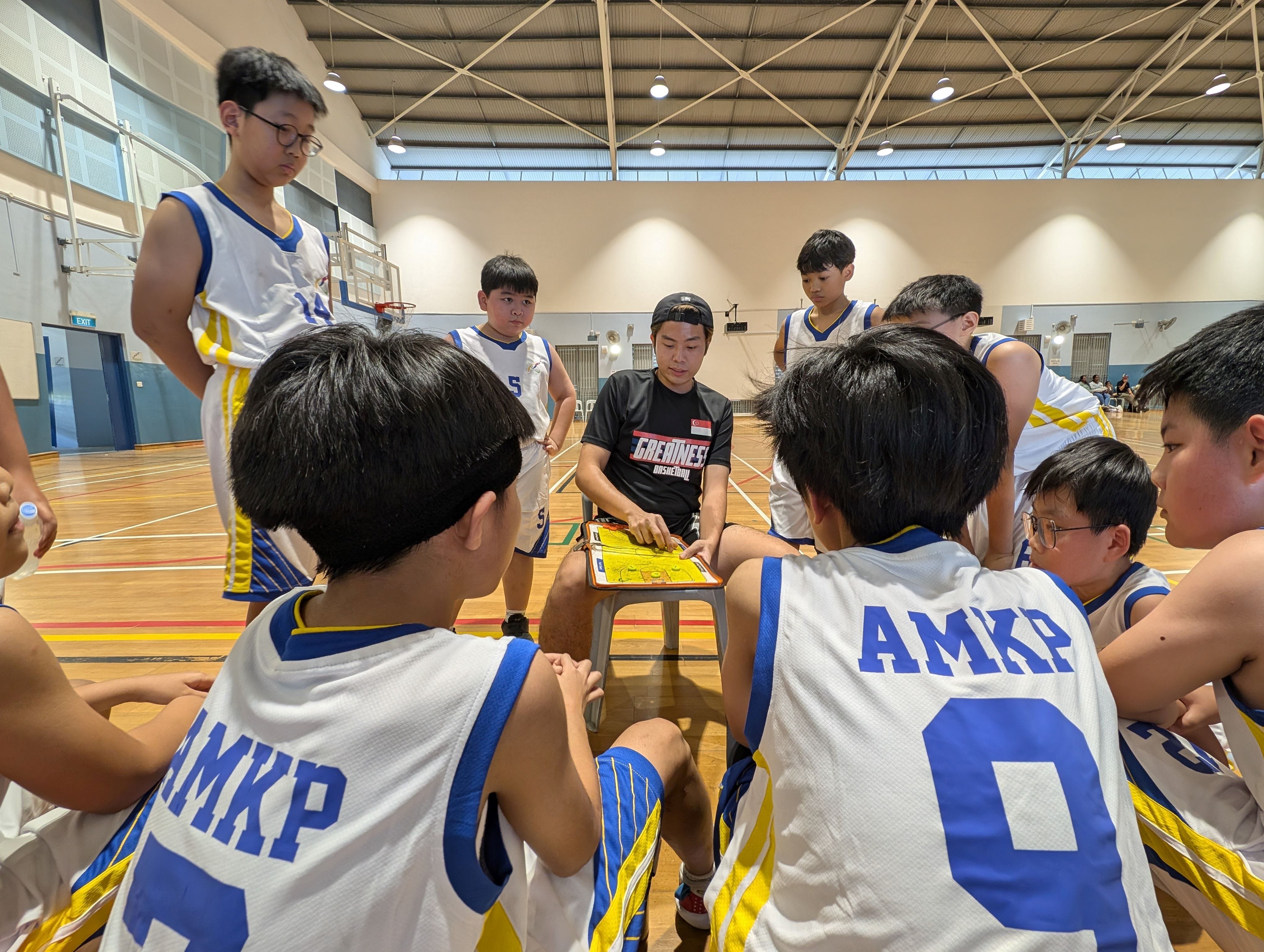 A coach draws plays on a clipboard while young basketball players in AMKP jerseys huddle around during a timeout.