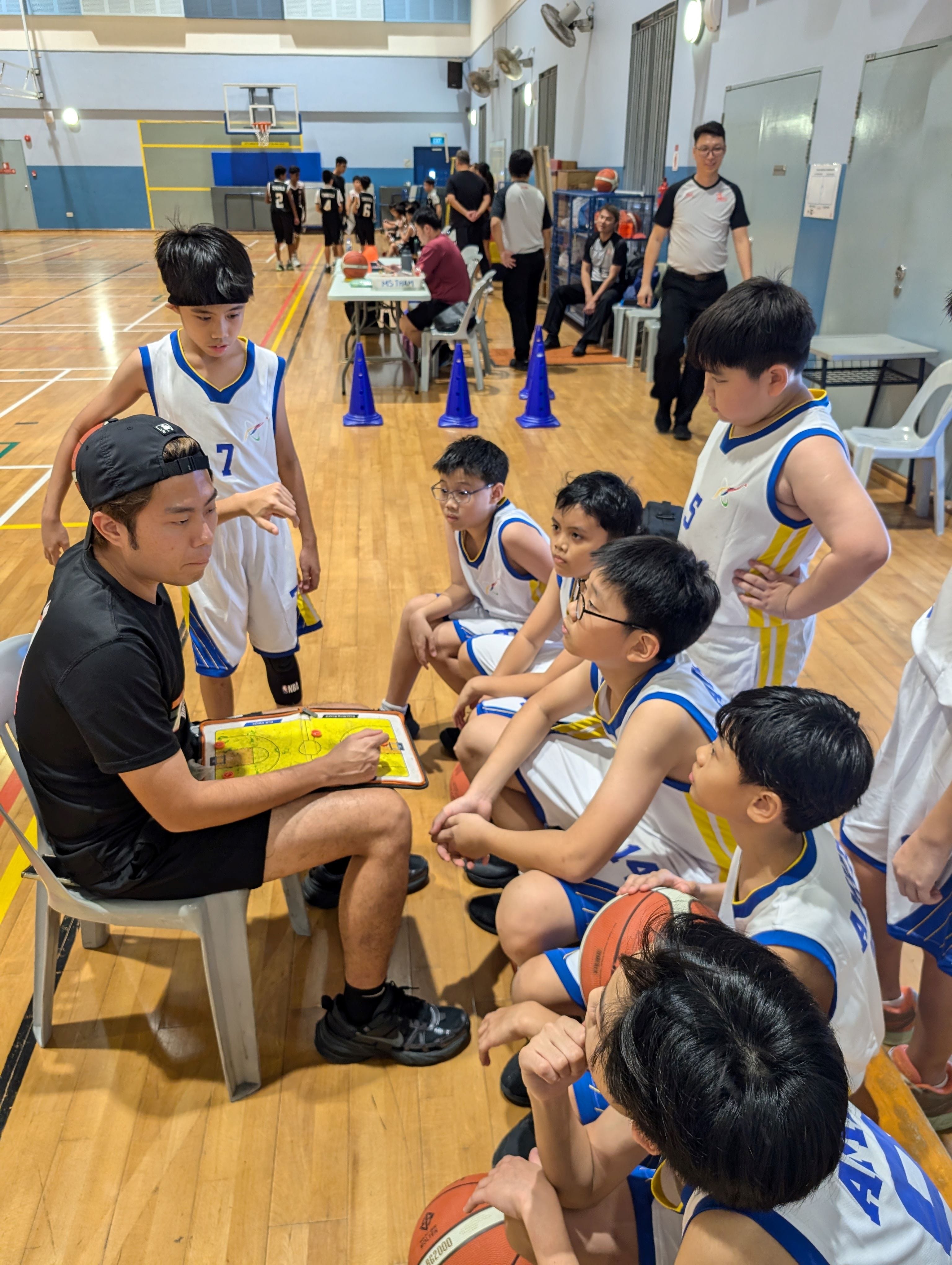 A coach shows a tactics board to young basketball players in white uniforms during a huddle in an indoor gym.
