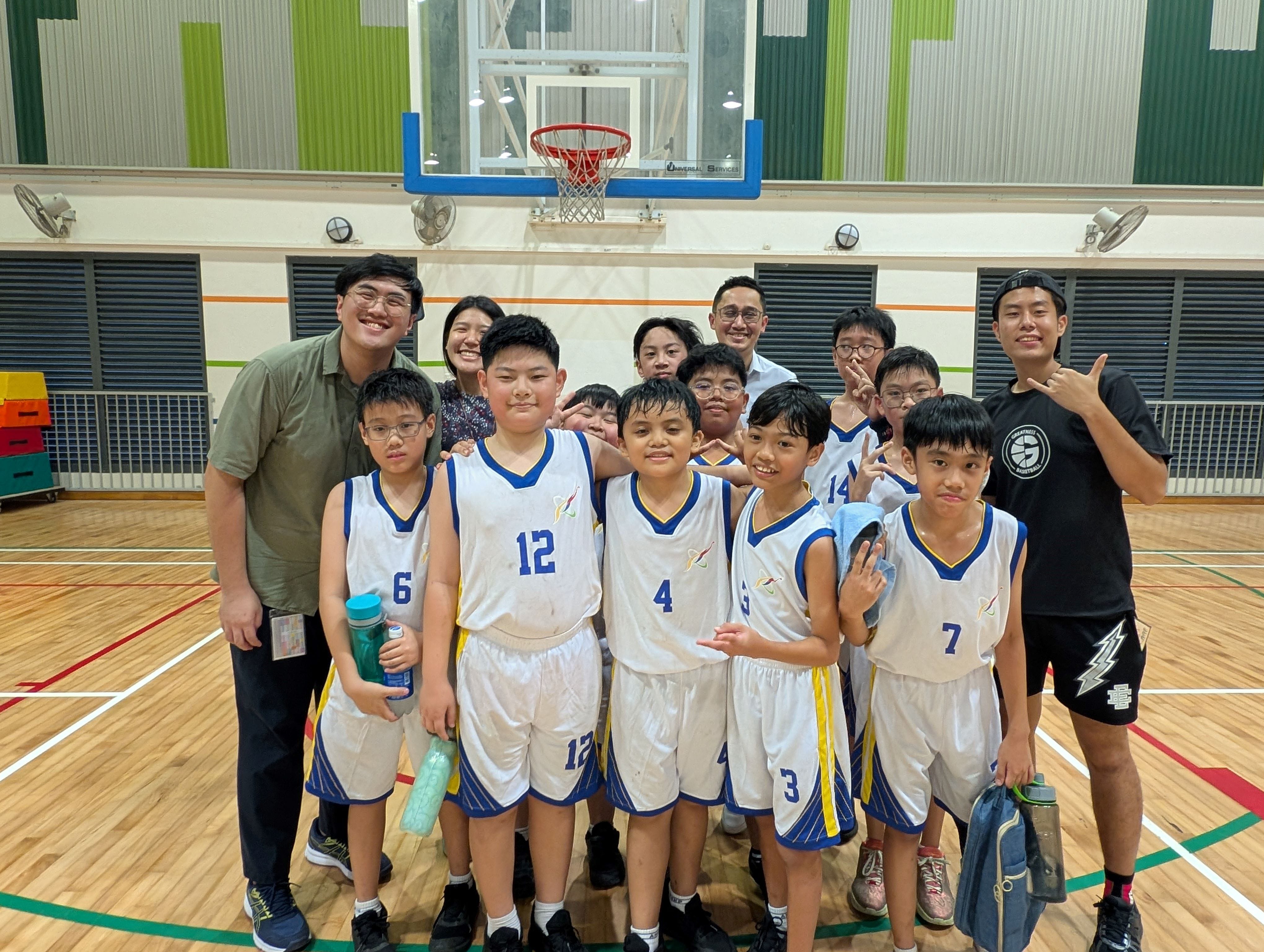 A youth basketball team in white and blue jerseys poses with coaches inside an indoor court with a hoop visible behind them.