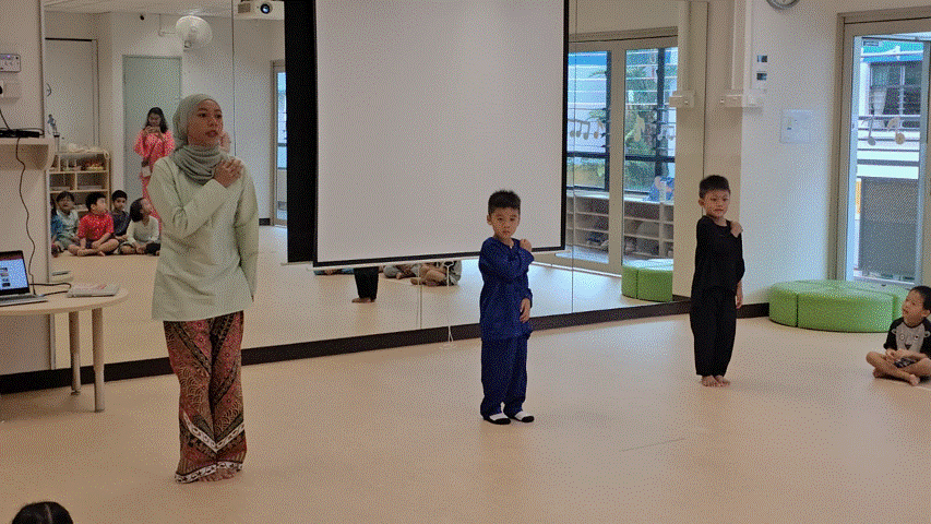 A woman in a hijab and two young boys stand performing in a bright classroom with a projection screen.