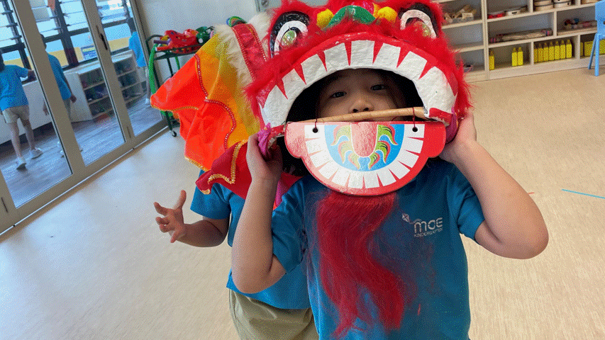 A young child holds a colorful red lion dance costume head over their face in a classroom.