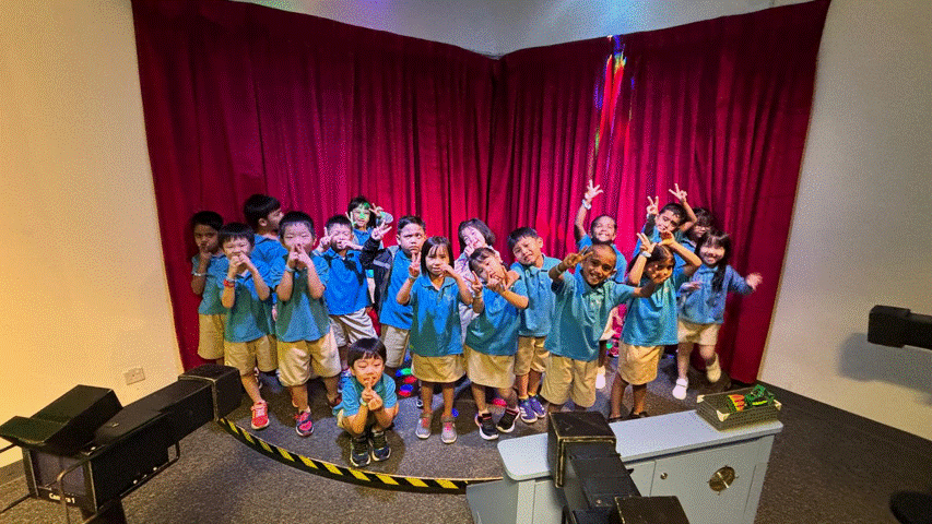 A group of young children in blue shirts and tan shorts pose cheerfully in front of a red curtain on a small stage.
