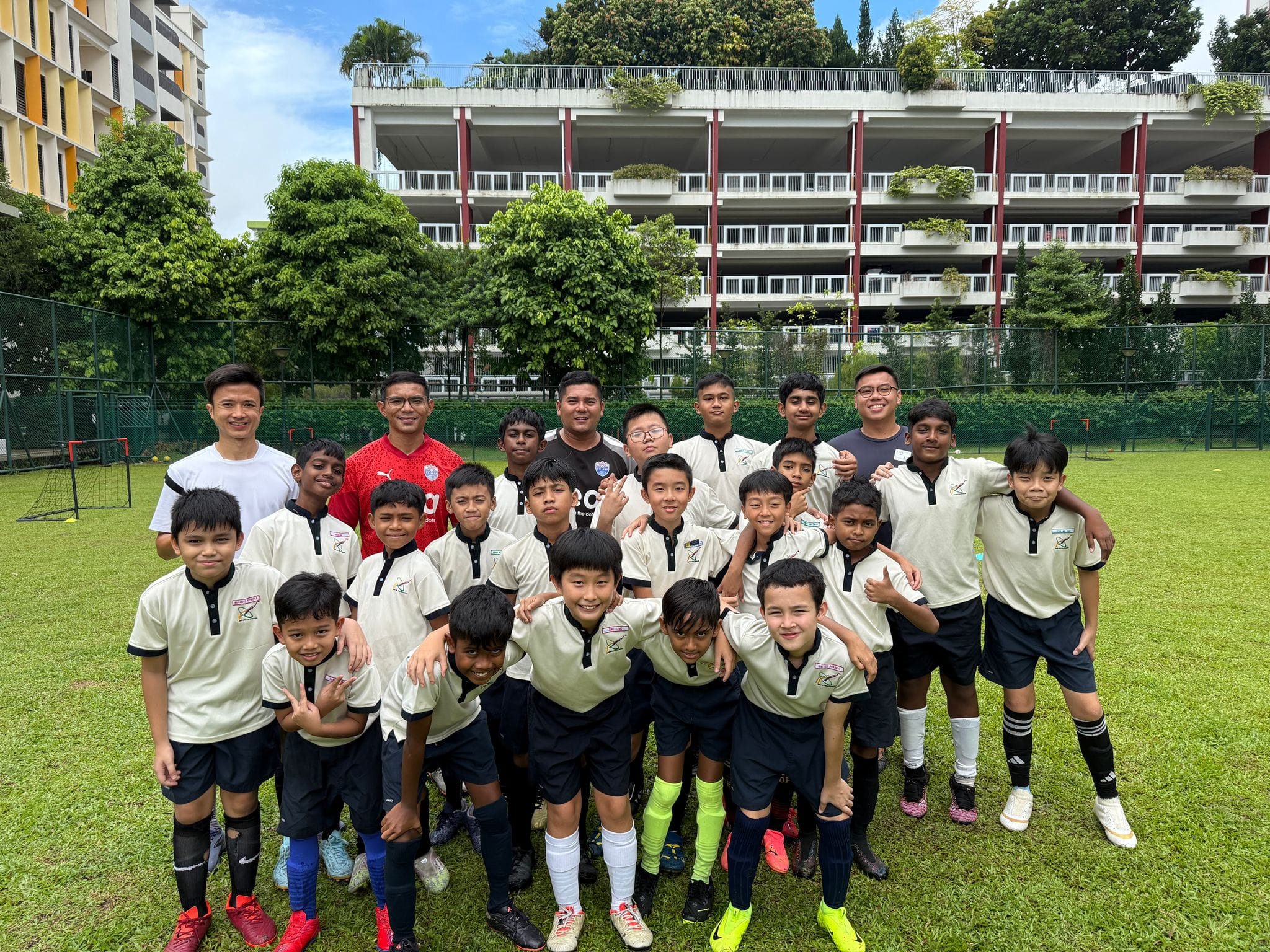 A youth soccer team in white and black uniforms poses with coaches on a green field in front of a multi-story building.