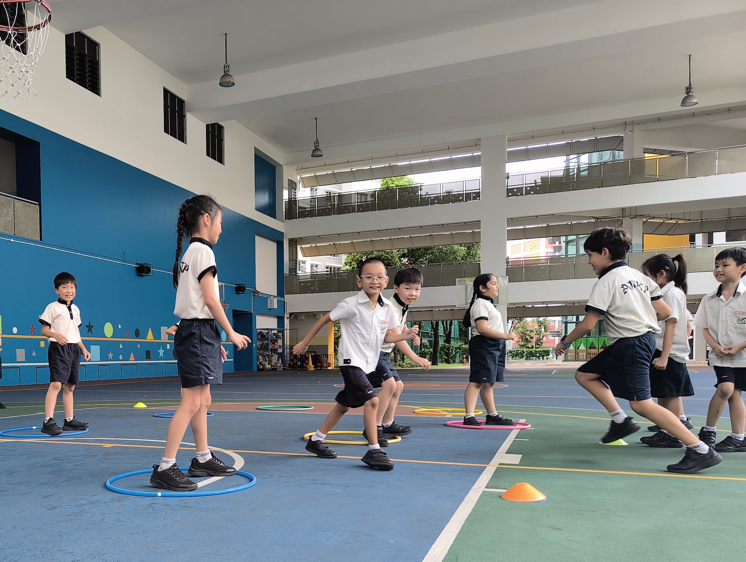 Children in school uniforms play an active game with colorful hoops and cones on an indoor sports court.