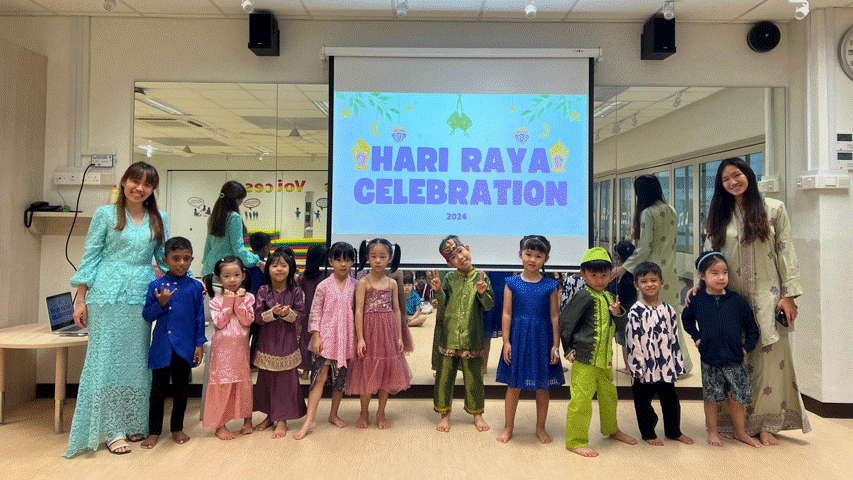 Children and two adults in traditional outfits pose in front of a screen displaying "Hari Raya Celebration 2024."