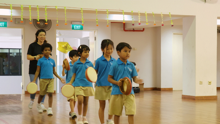 Young children in blue uniforms carry tambourines while walking in a line inside a decorated hall, led by a teacher.