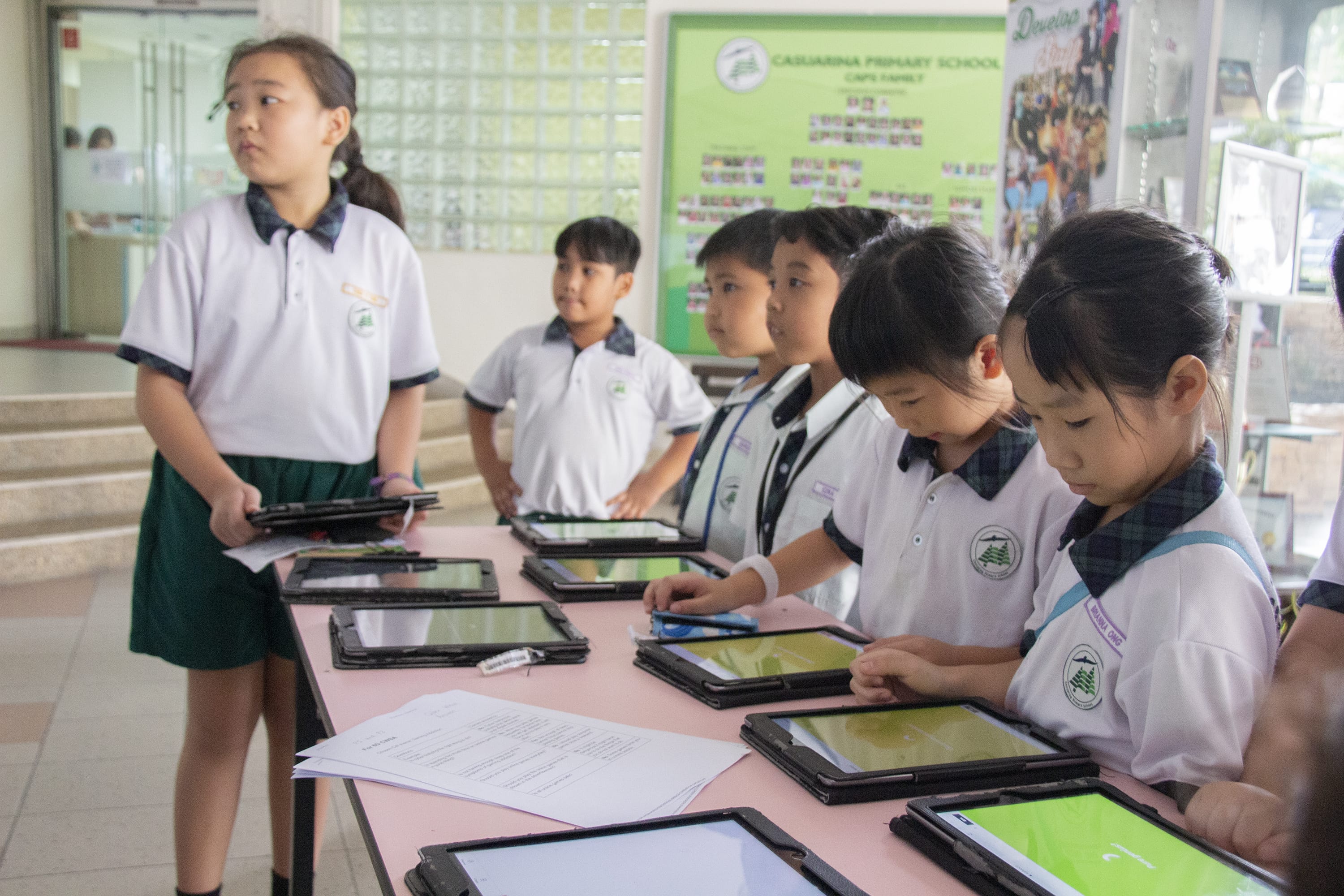 Schoolchildren in uniforms stand at a table with tablets, one girl holding a tablet and looking left.