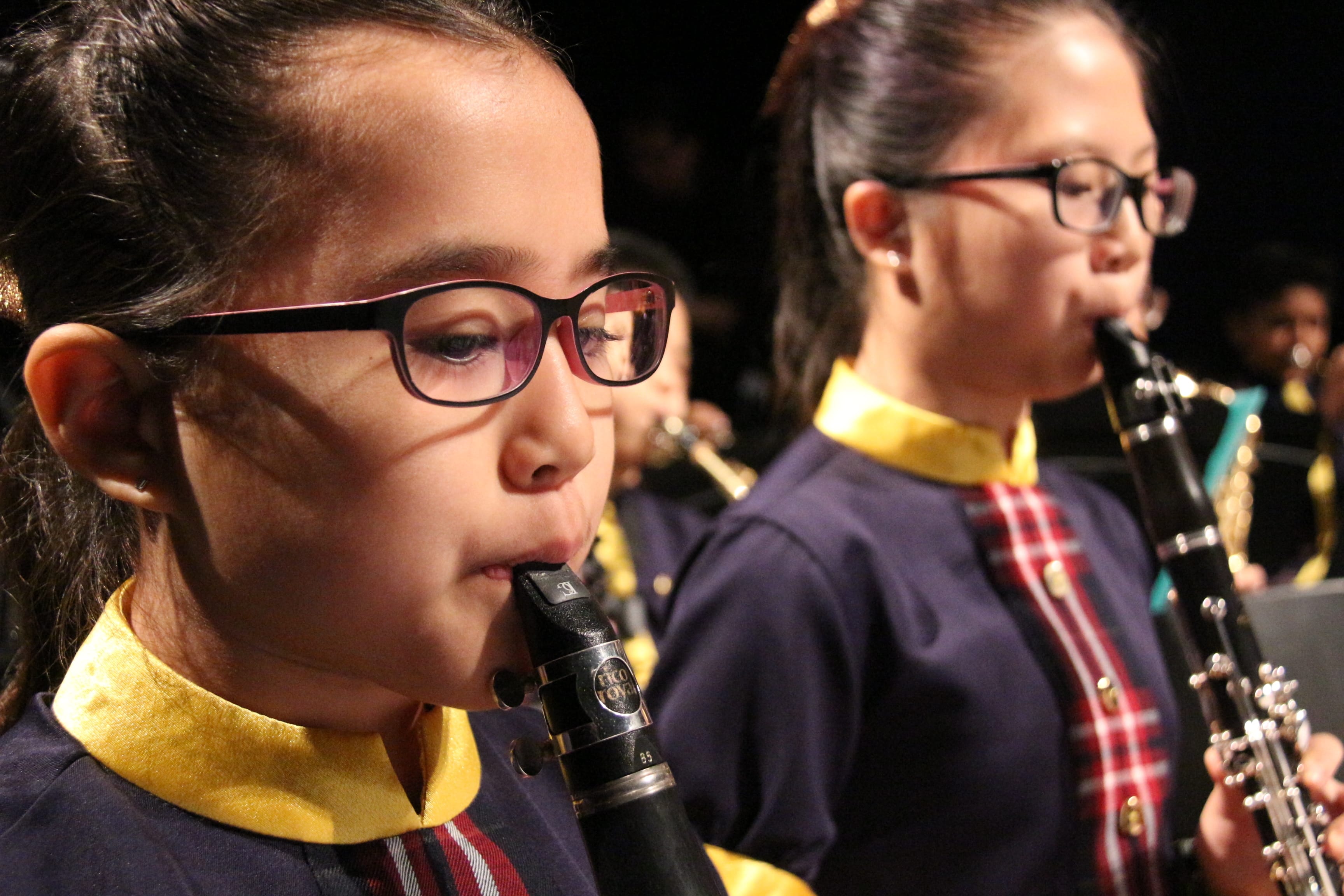 Two young girls wearing glasses and school uniforms play clarinets in a youth band performance.