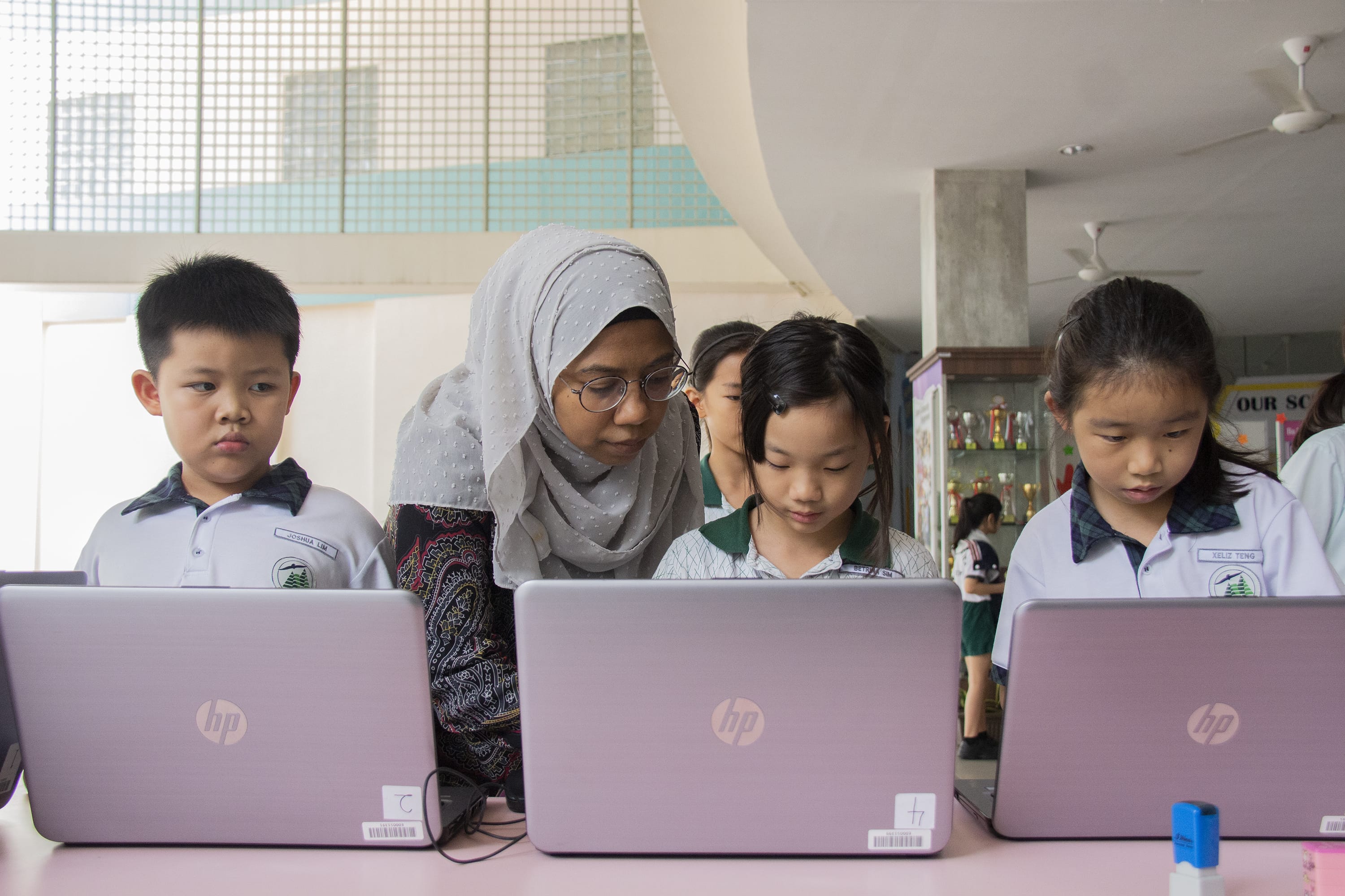 A teacher wearing a hijab leans over to assist young students working on HP laptops in a classroom.