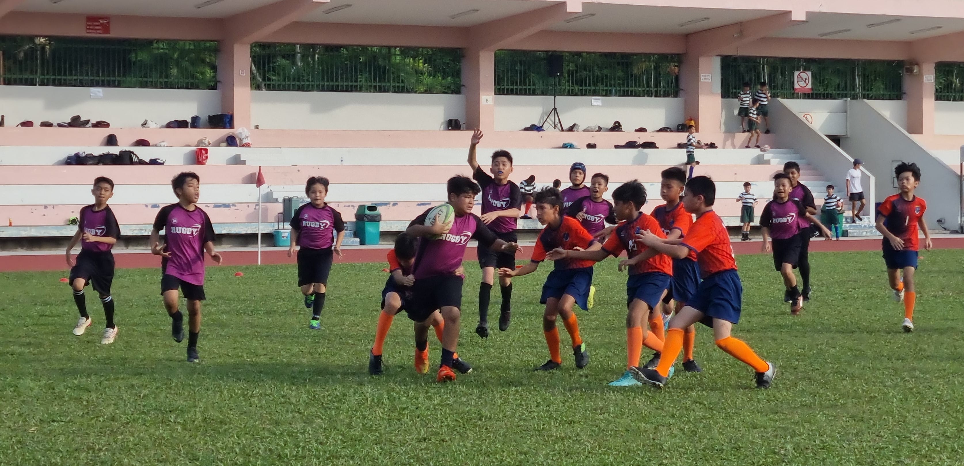 Young boys in purple and orange jerseys play rugby on a grass field with stadium bleachers in the background.