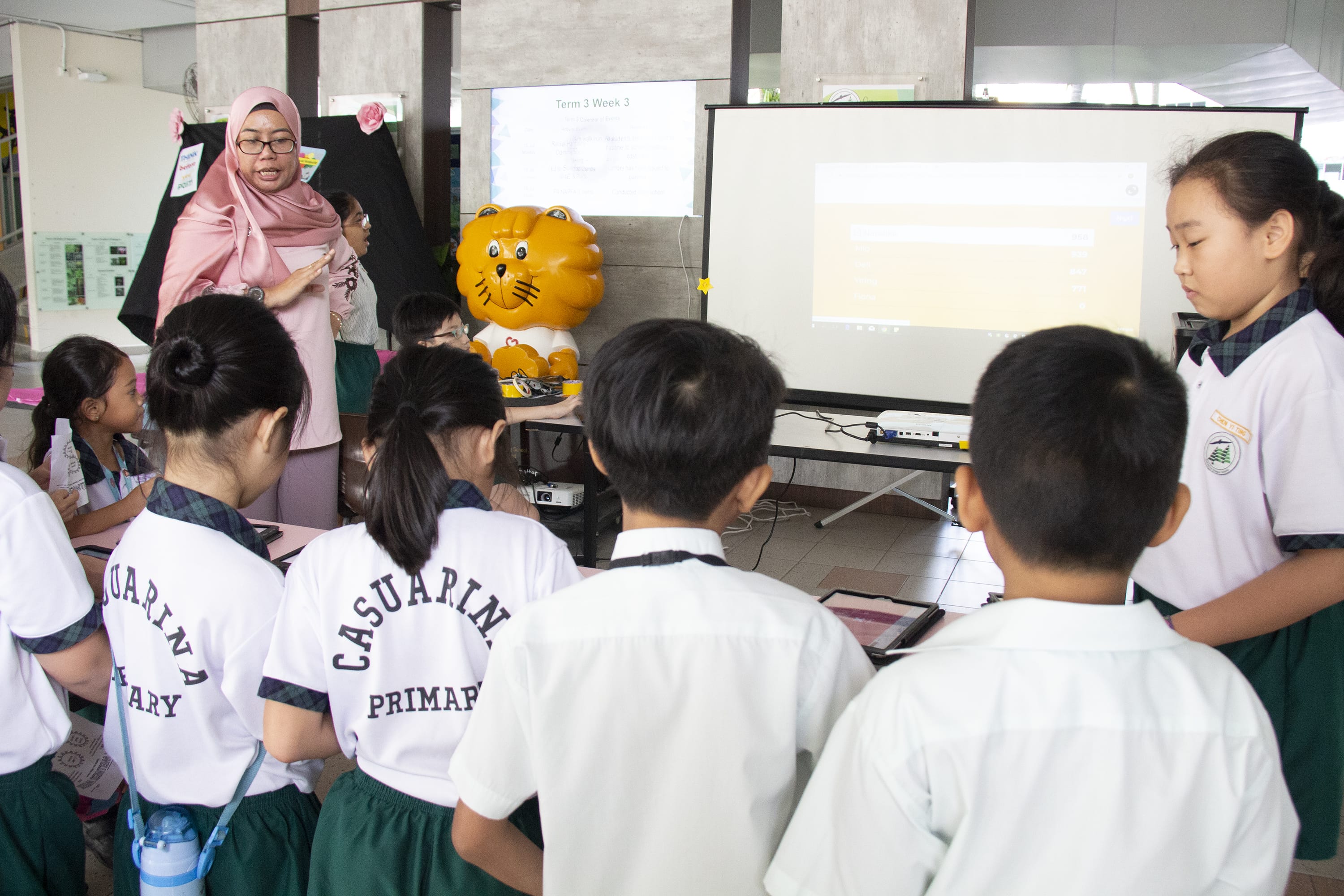 Teacher in pink hijab speaks to uniformed students as a projector screen shows a lesson in a classroom.
