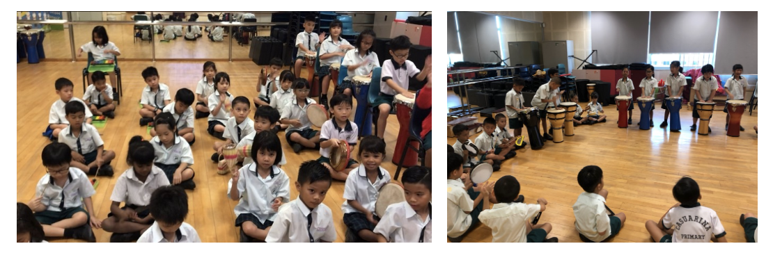 Young students in white uniforms sit on a wooden floor playing hand drums and percussion instruments in a school hall.