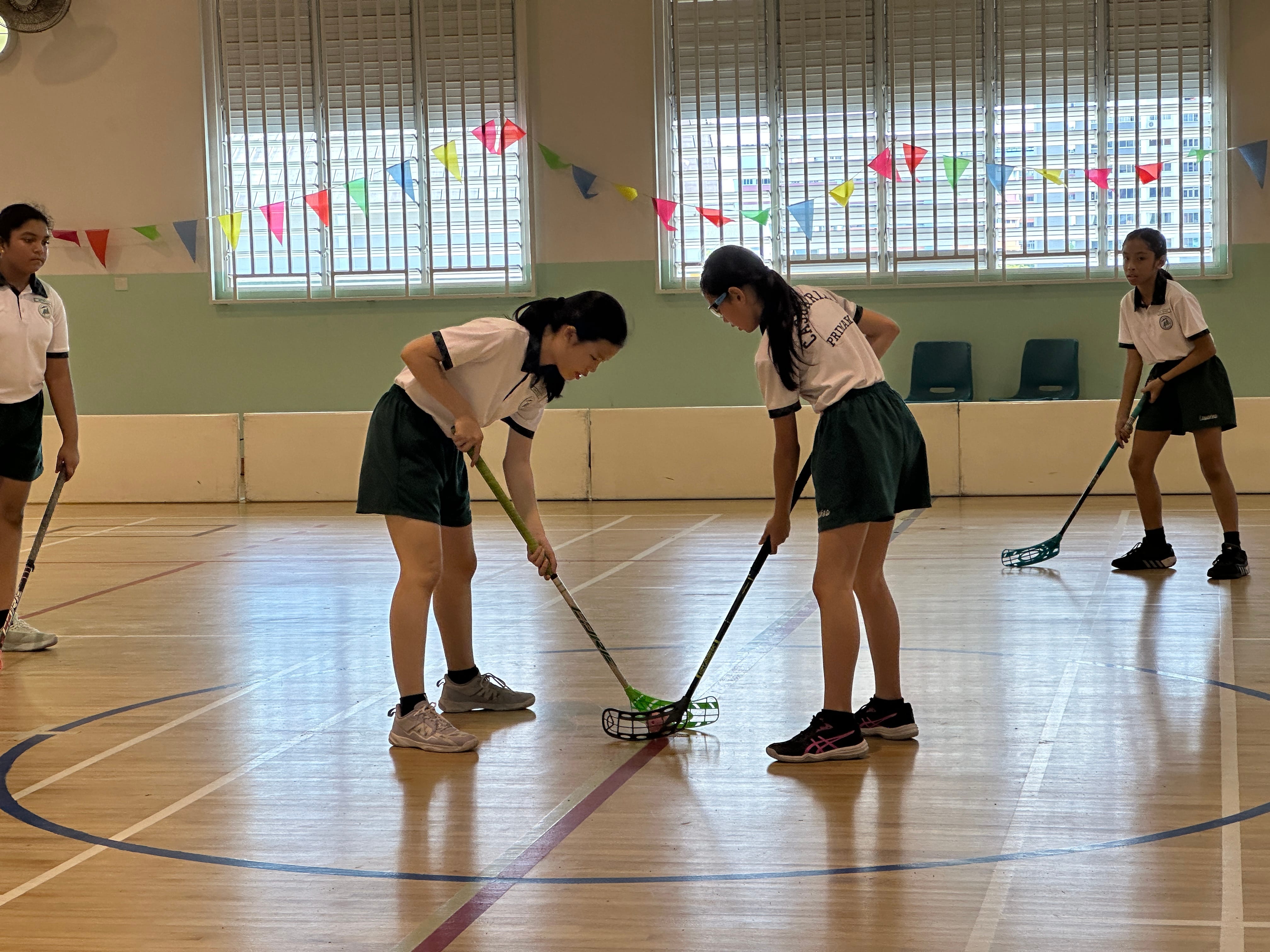 Four girls in school uniforms play floorball with sticks in an indoor gymnasium decorated with colorful bunting.